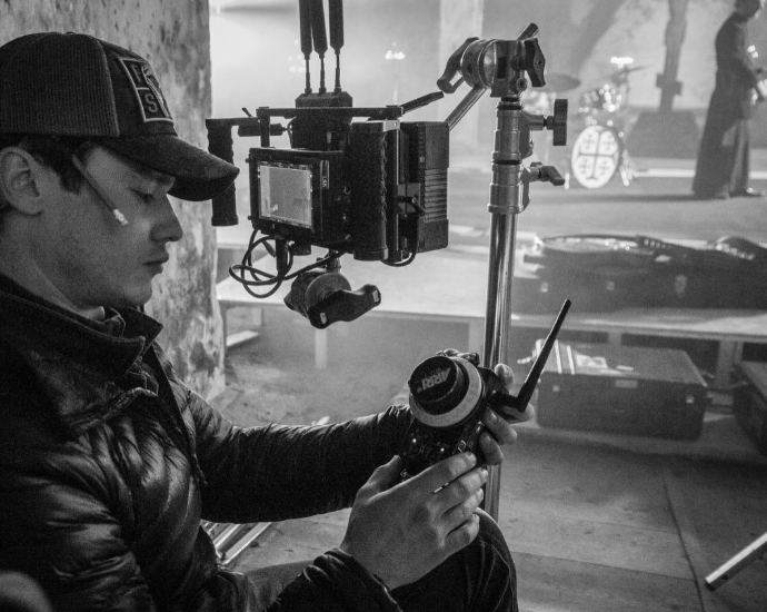 A person wearing a cap and jacket operating a camera on a steadicam rig, filming in an indoor location.