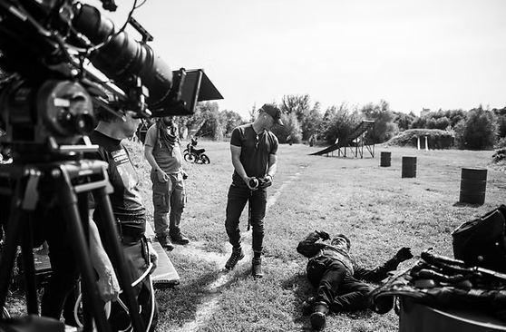 Filming of a crime scene with a person lying on the ground and crew members setting up equipment in an outdoor park.