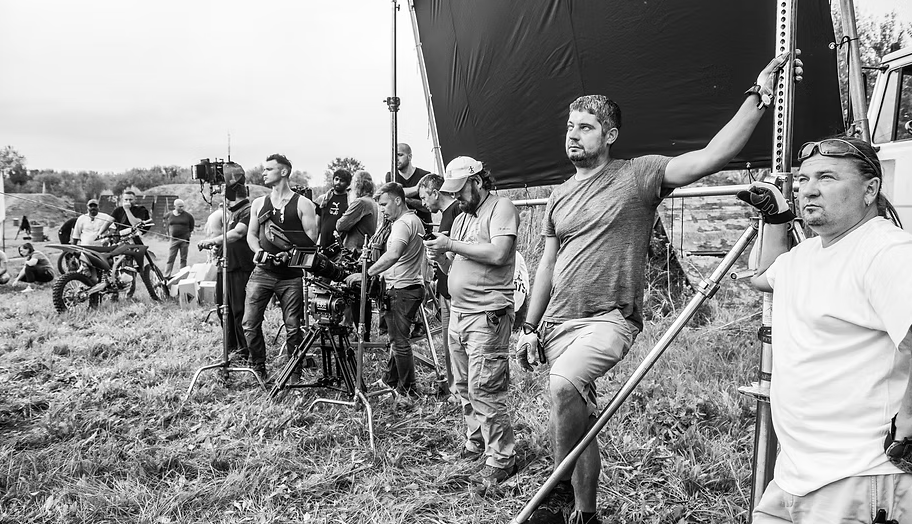Behind-the-scenes photo of a film shoot outdoors, with crew members and actors, some standing near camera and equipment, with a field and trees in the background.