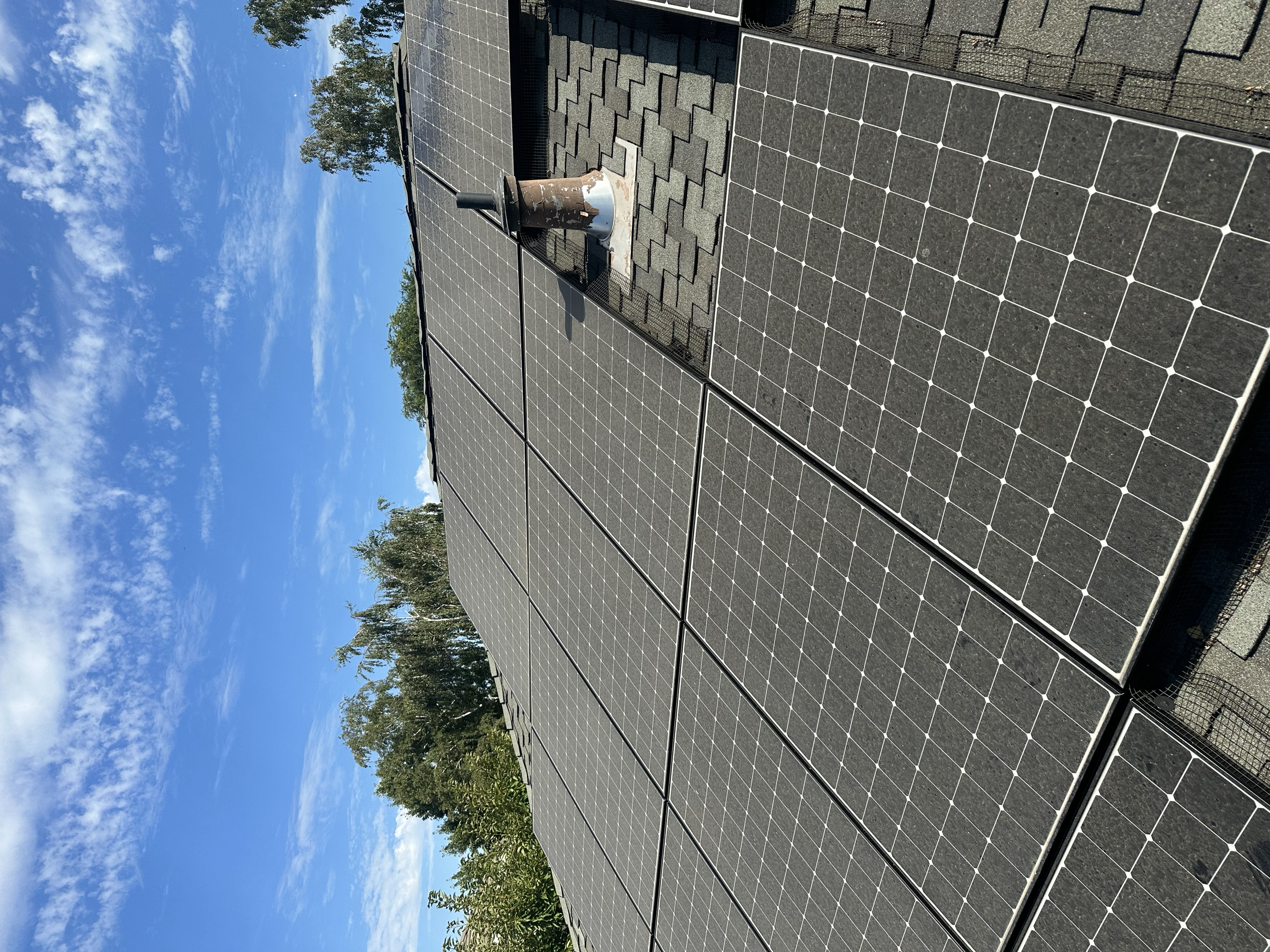 Solar panels installed on a rooftop under a partly cloudy blue sky, with trees in the background.