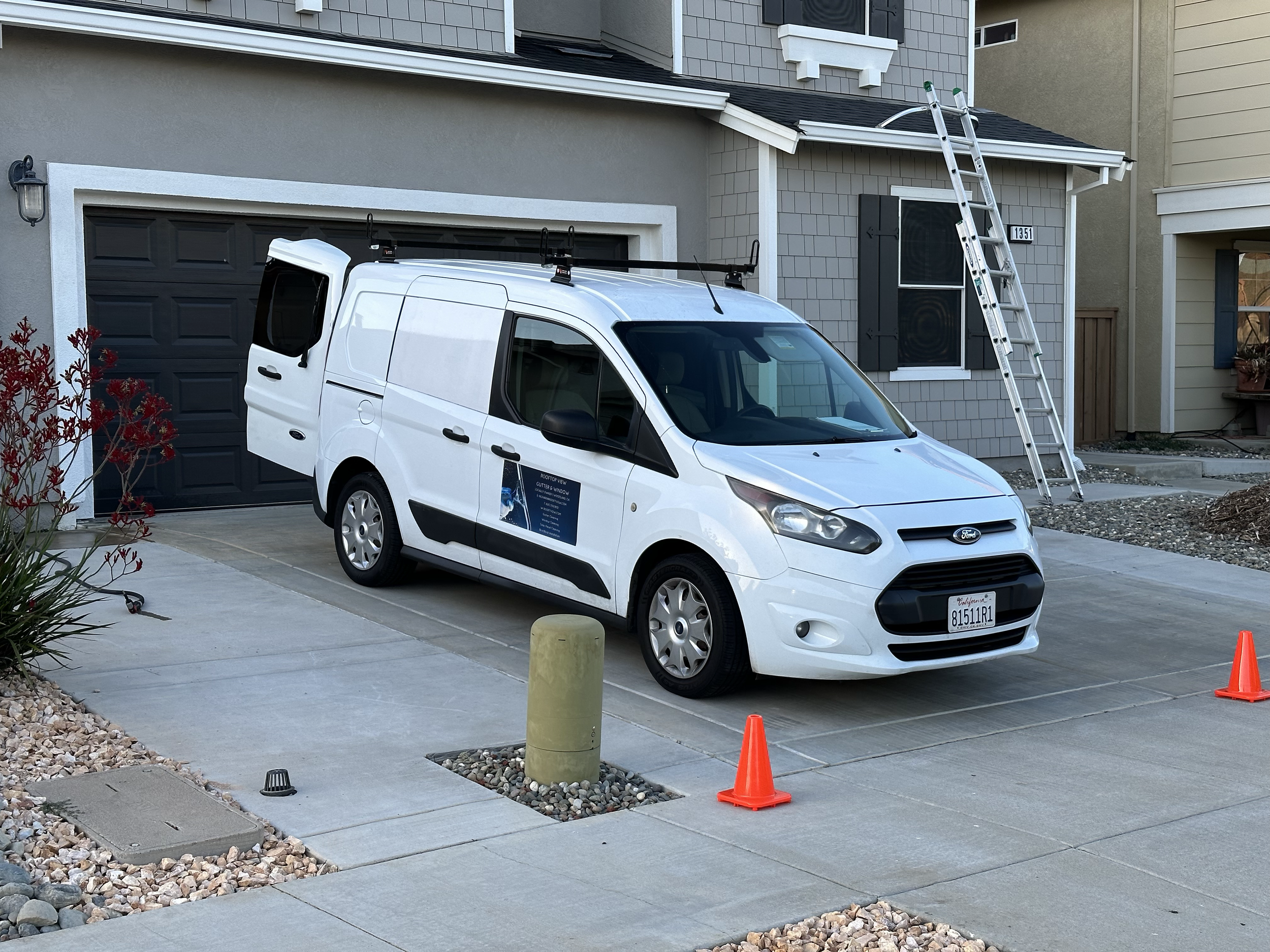 White Ford transit Connect van parked in driveway with orange cones around it, ladder leaning against house, and a door open with tools inside.