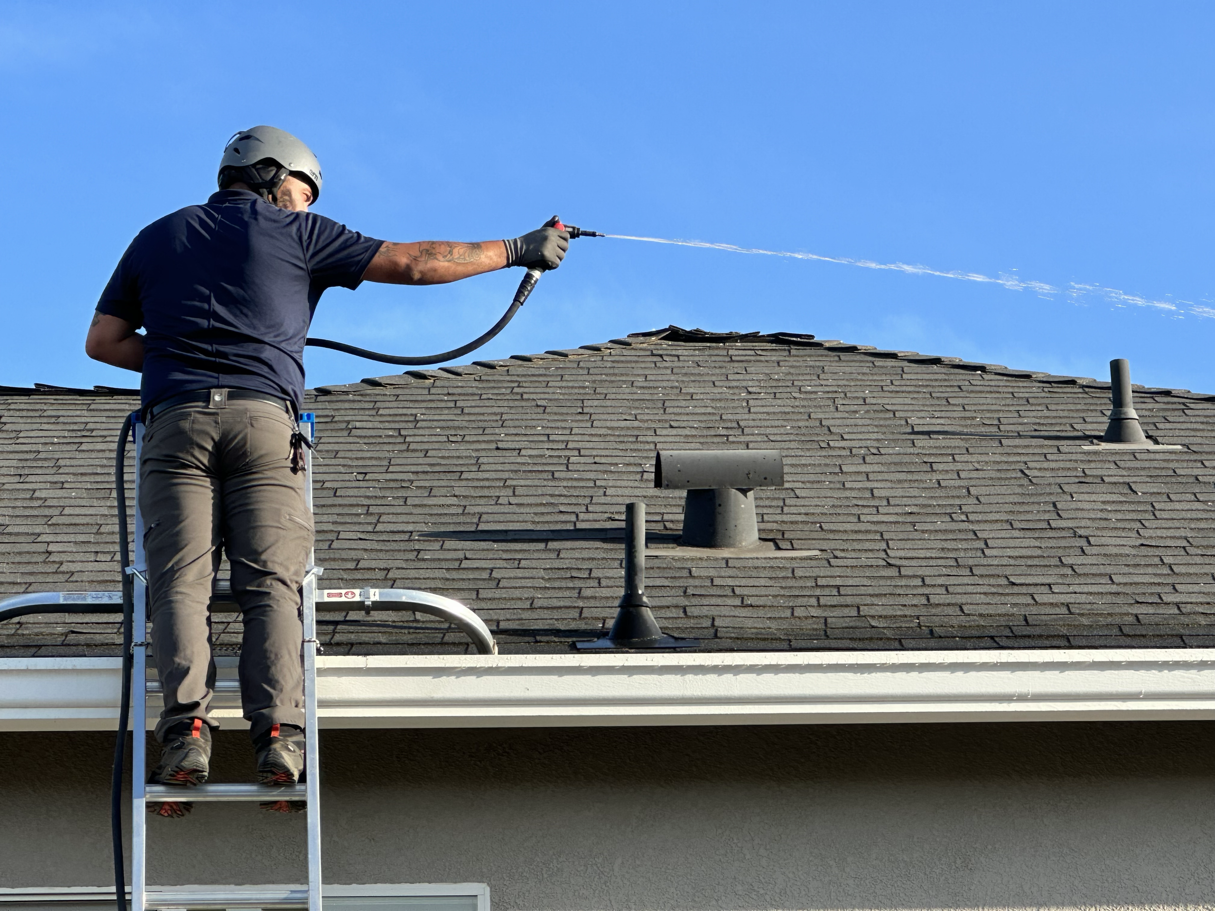 A person wearing a helmet and gloves standing on a ladder, pressure washing the roof of a house under a clear blue sky.