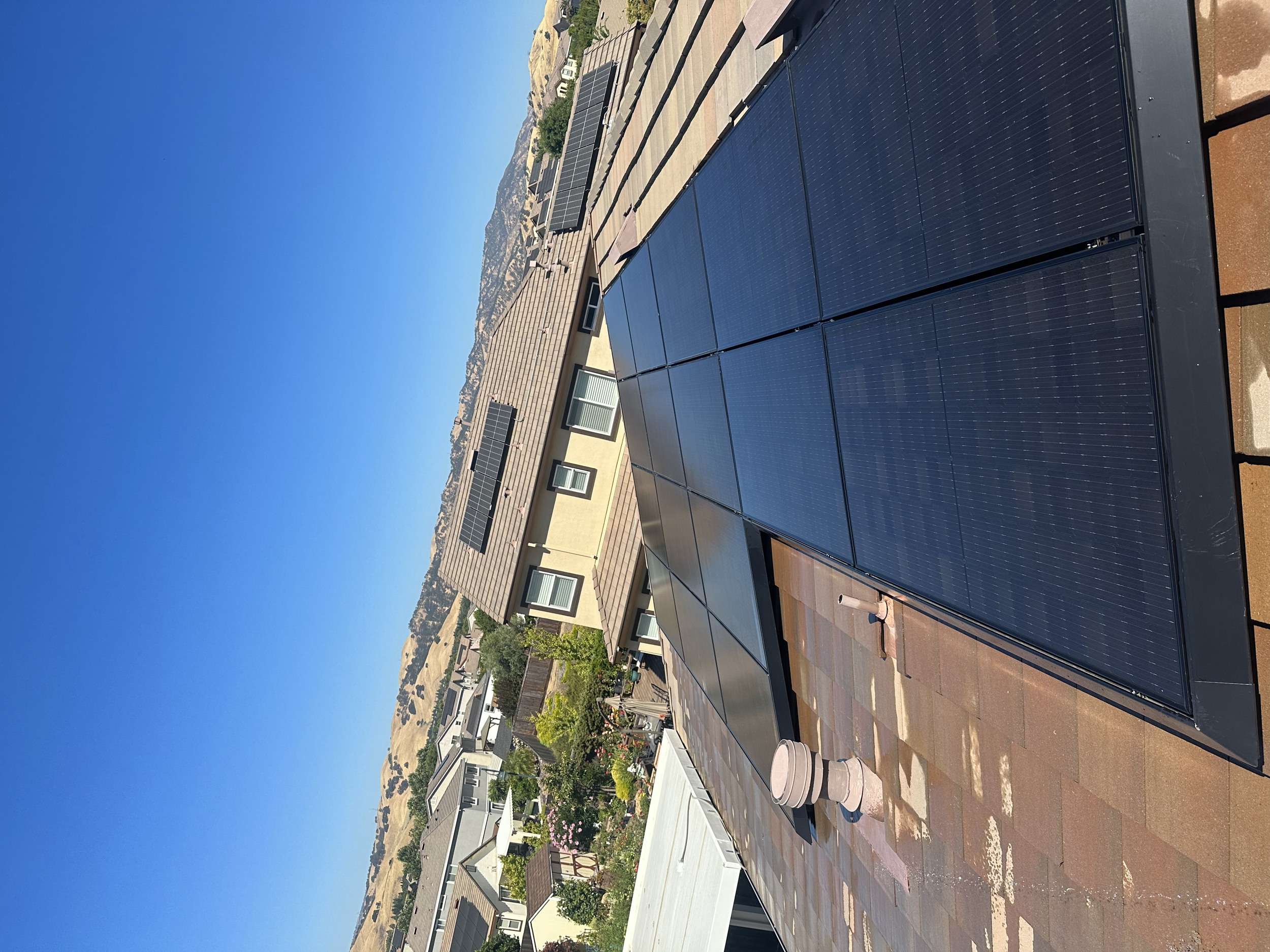Solar panels installed on a rooftop under a clear blue sky, with neighboring houses and hilly terrain in the background.