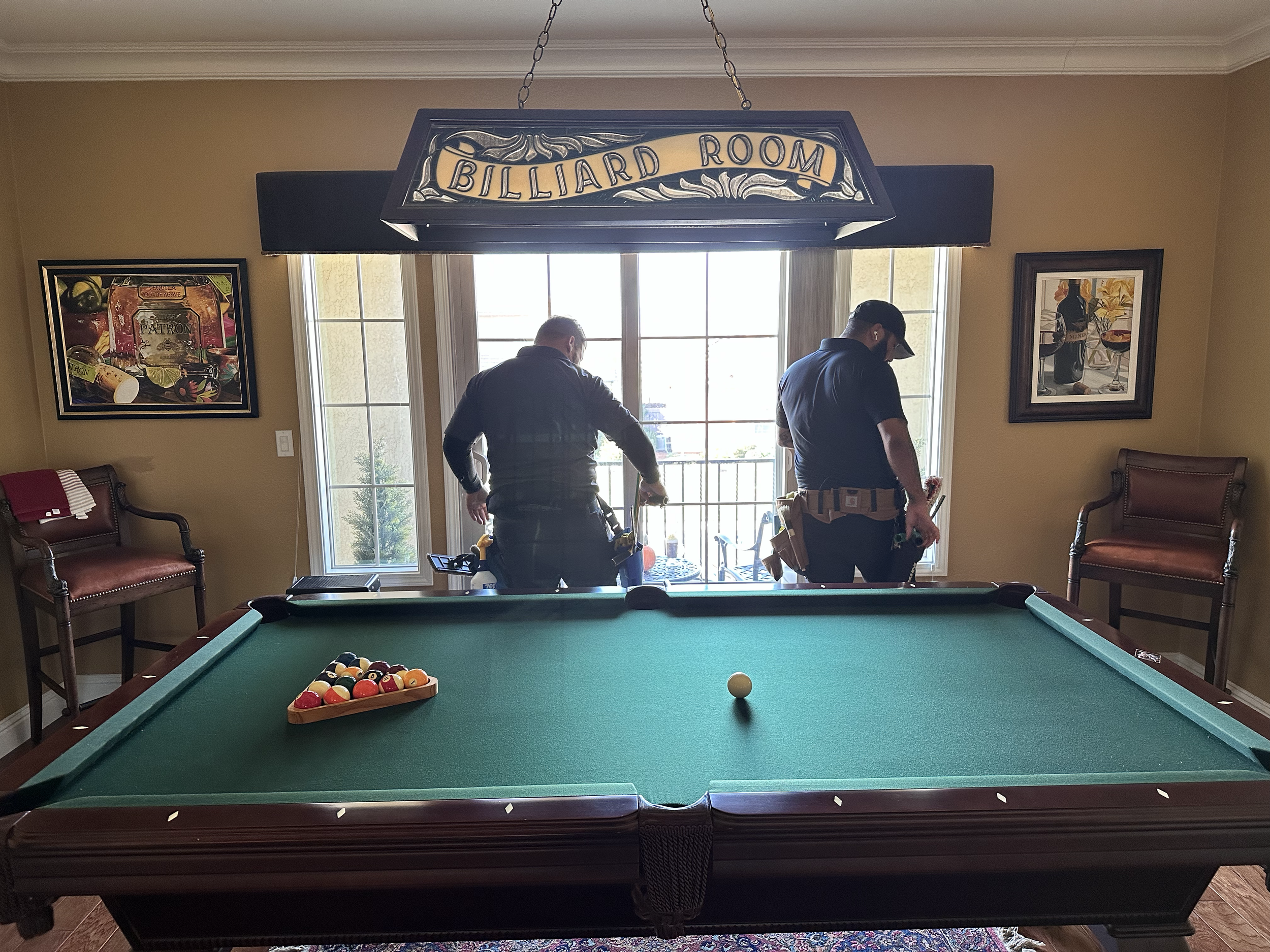 Two workers in billiard room setting up or maintaining pool table, with a pool cue on the table and pool balls racked in triangle, standing near large window with patio furniture outside, under a hanging sign that says 'Billiard Room'.