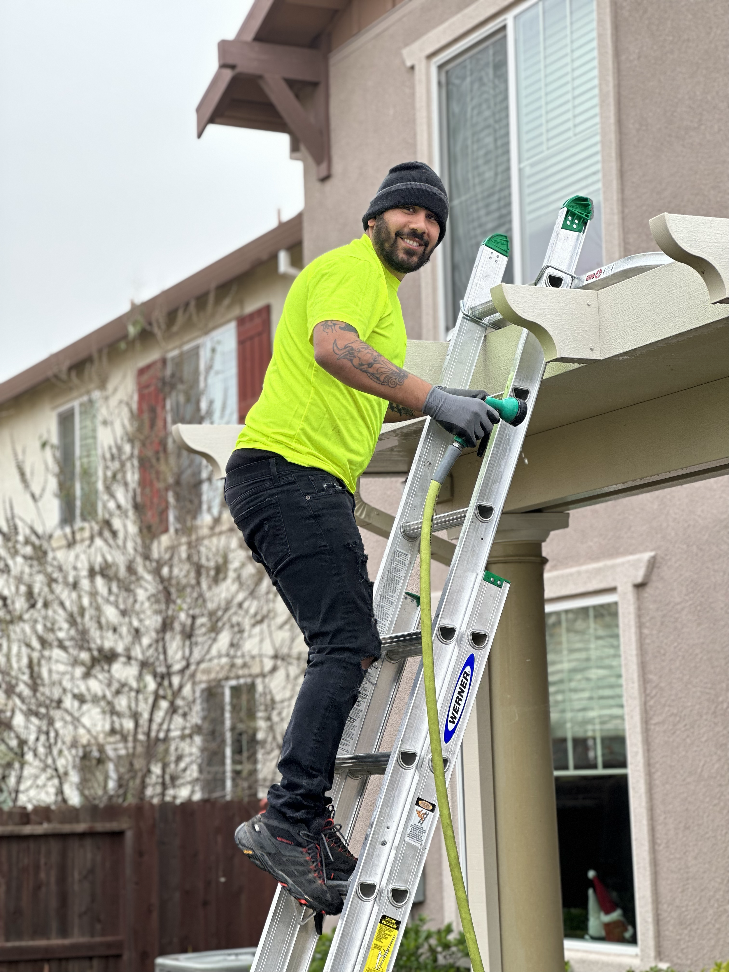A man in a neon yellow shirt and black pants is smiling on a ladder outside a house, working on the roof with tools.