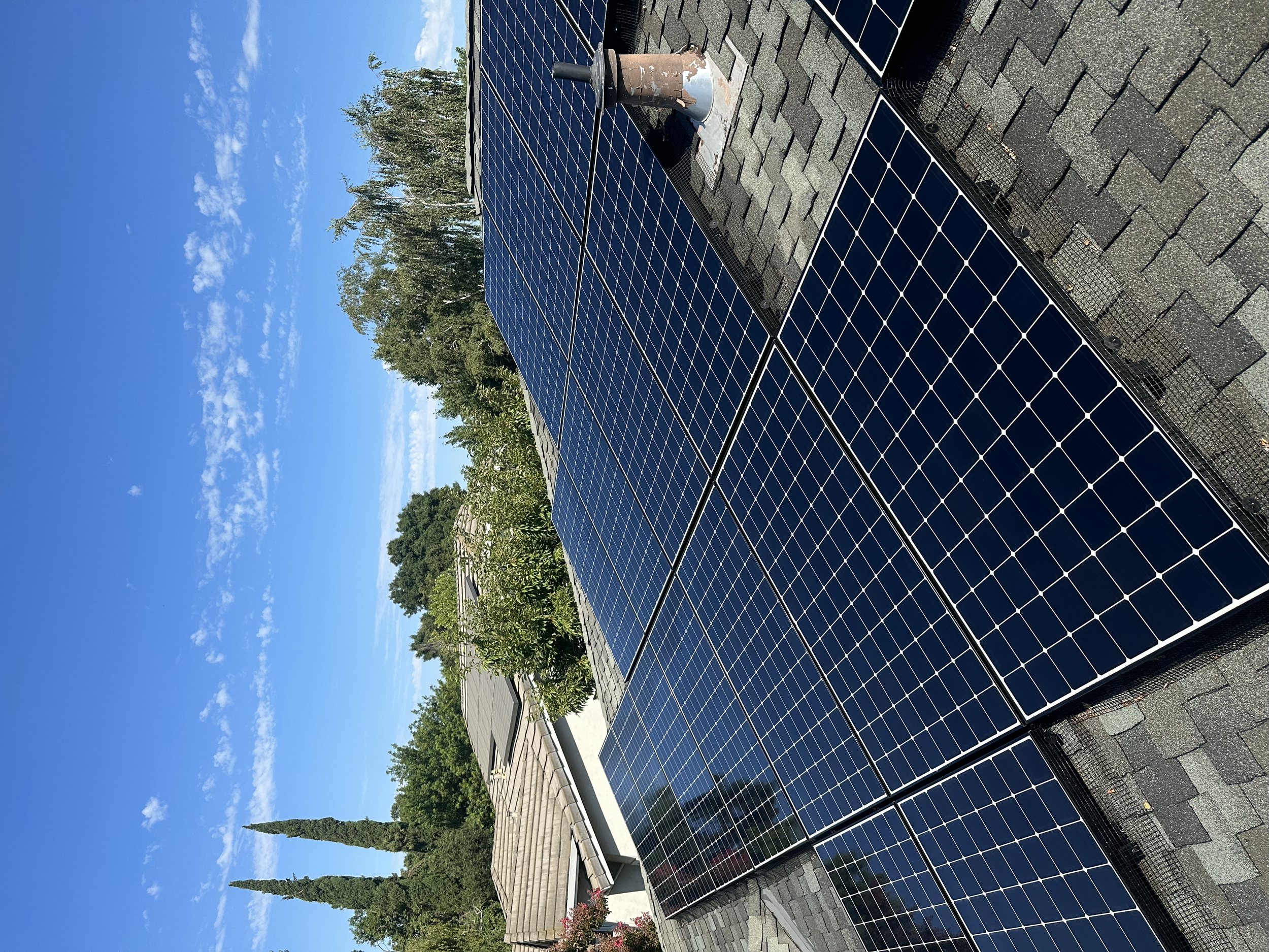 Solar panels installed on a rooftop under a clear blue sky with some scattered clouds, greenery, and trees in the background.