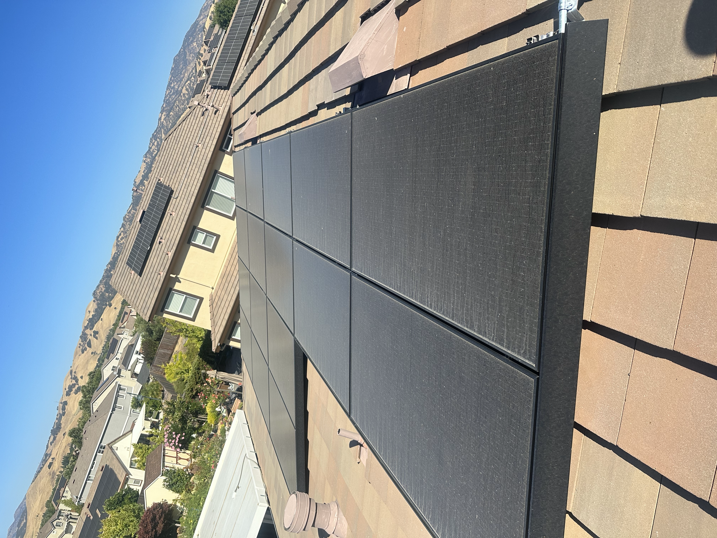 Solar panels installed on the roof of a house with neighboring houses and hills in the background.