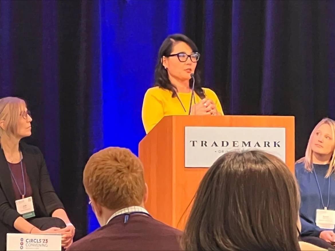 Women at a conference, one woman in a yellow top stands at a podium labeled 'TRADEMARK' speaking into a microphone, with two seated women to her left and right, all wearing conference badges.