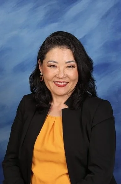 A woman with dark hair, wearing a black blazer and yellow top, smiling against a blue cloudy background.
