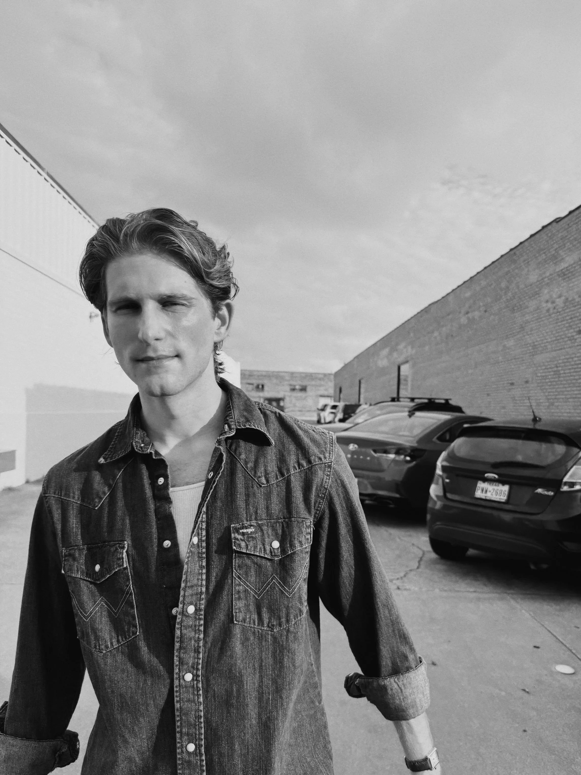 Black and white photo of a young man with wavy hair in a denim jacket standing in a parking lot next to a brick wall and several parked cars, with a partly cloudy sky in the background.