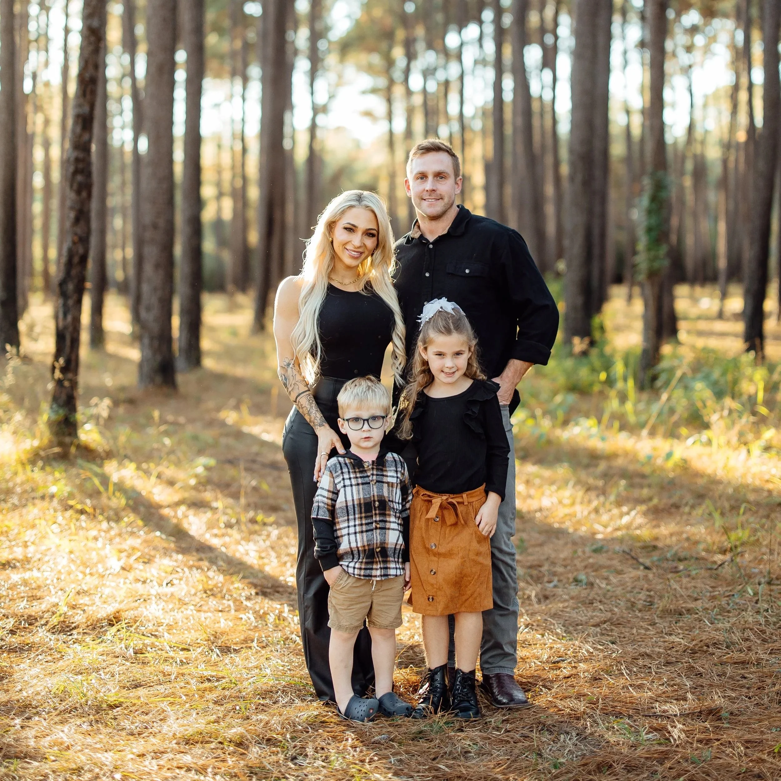 A family of four standing in a sunlit forest, smiling at the camera.