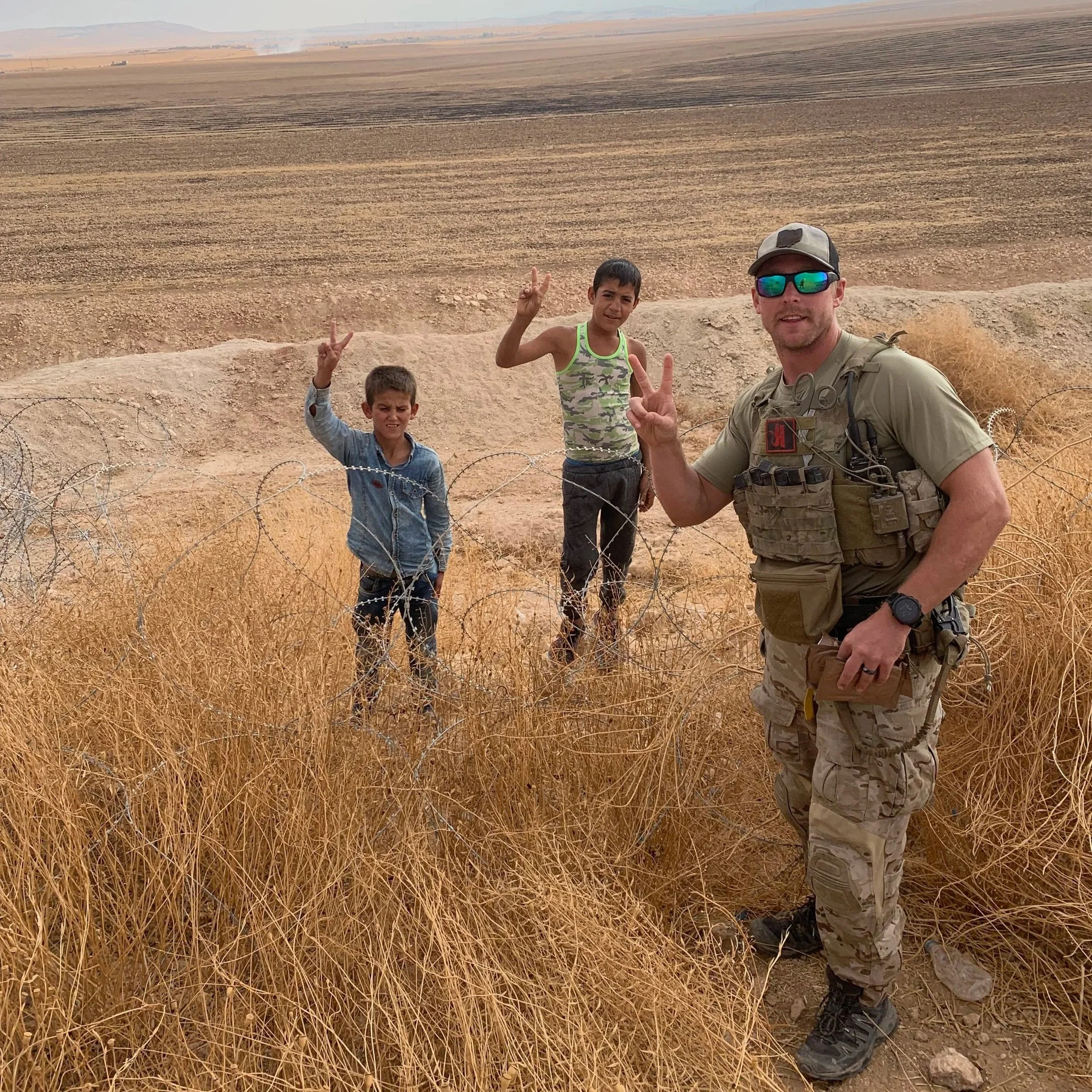 A soldier and two boys posing behind a barbed wire fence in a dry, barren landscape, making peace signs.