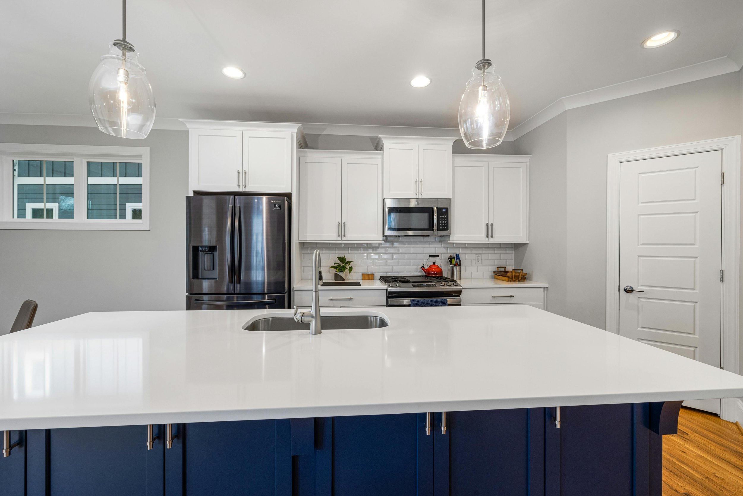 Modern kitchen with white cabinetry, stainless steel refrigerator, microwave, and stove, a white island with a sink, hardwood flooring, and pendant lights.