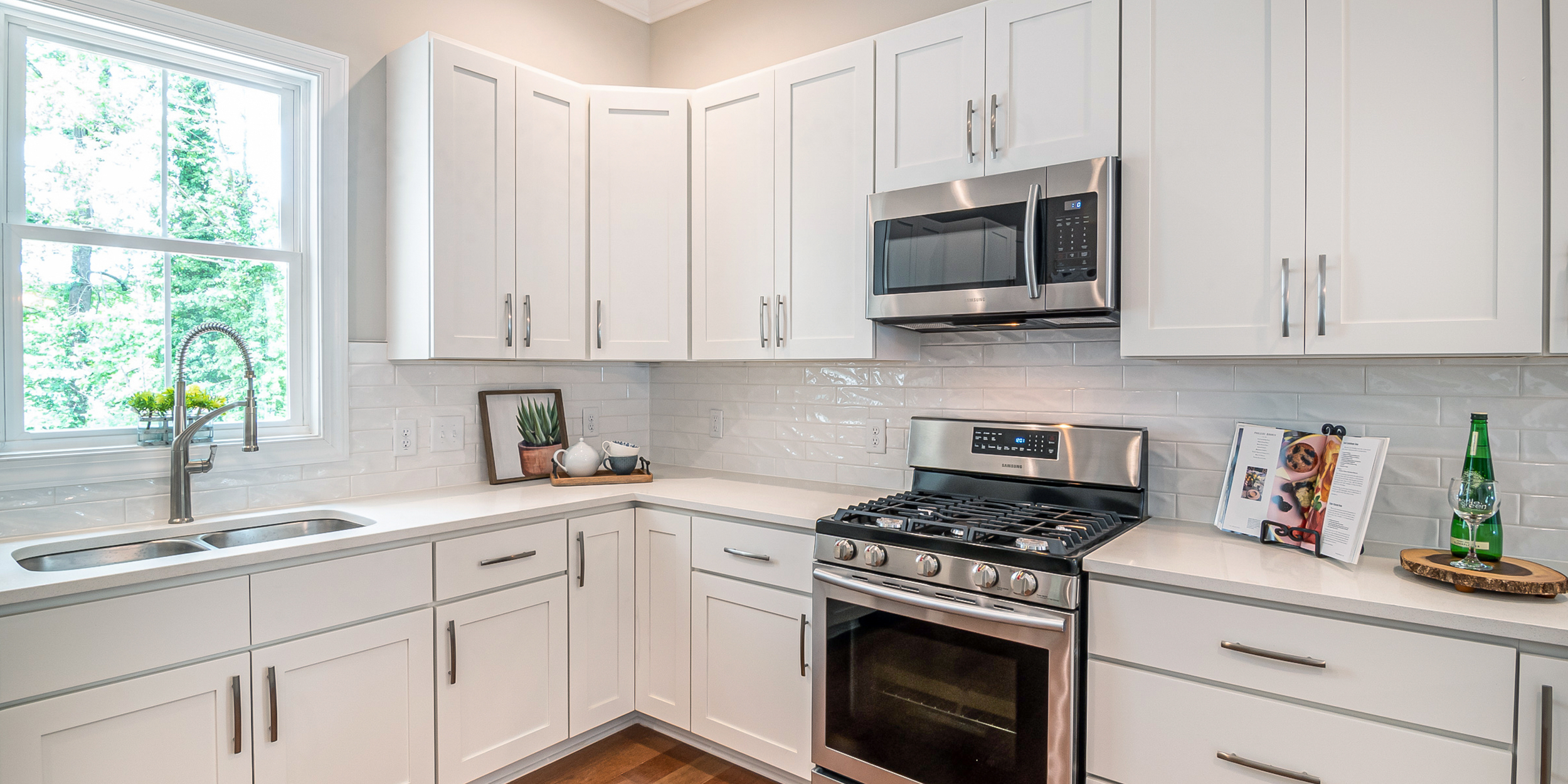 Modern kitchen with white cabinets, stainless steel microwave and oven, white countertops, a window above the sink, plants, a tray with a teapot and cups, an open cookbook with a plate of food, and a bottle of sparkling water on a wooden tray.
