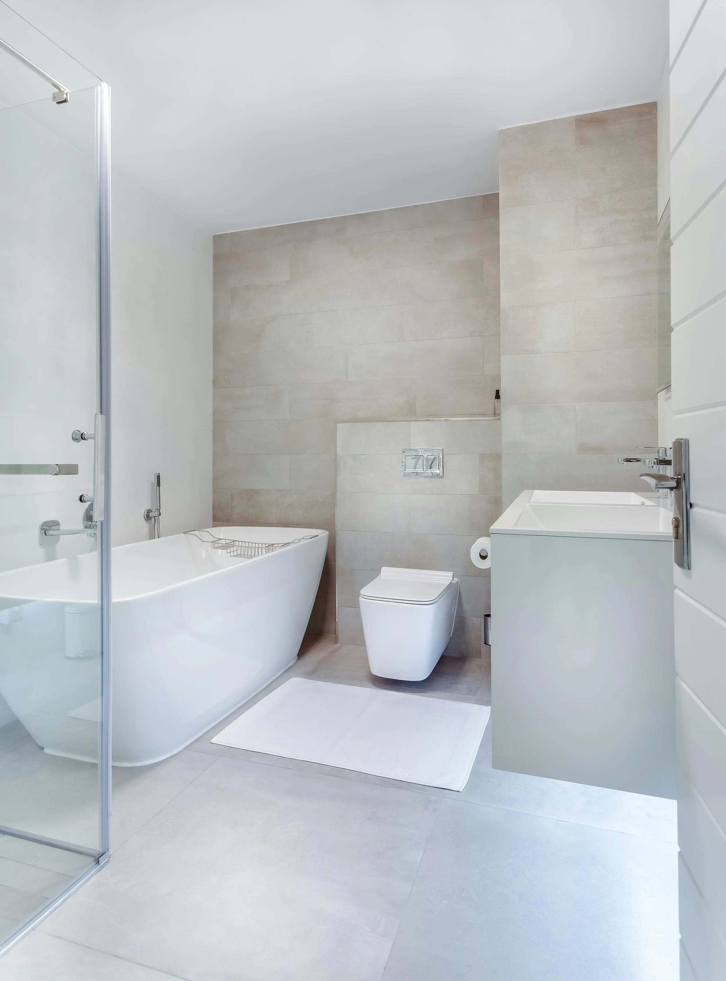 Modern bathroom with a white bathtub, toilet, gray tiled walls, white vanity, and glass shower enclosure.