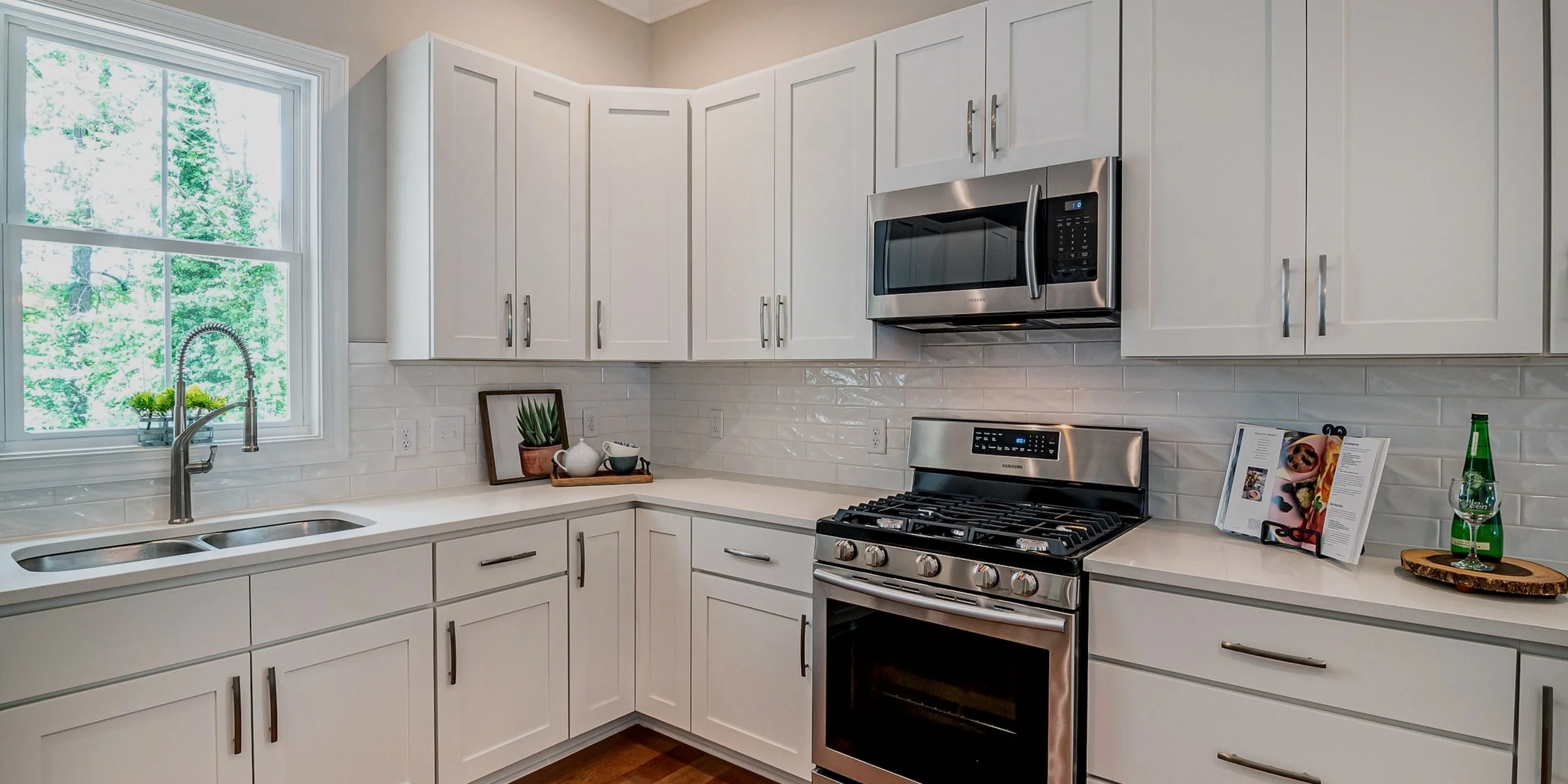A modern kitchen with white cabinets, a stainless steel microwave and stove, a white countertop, and a window with green trees outside. Decor includes potted plants, a tray with cups, open cookbook, and a green glass bottle with a glass on a wooden tray.