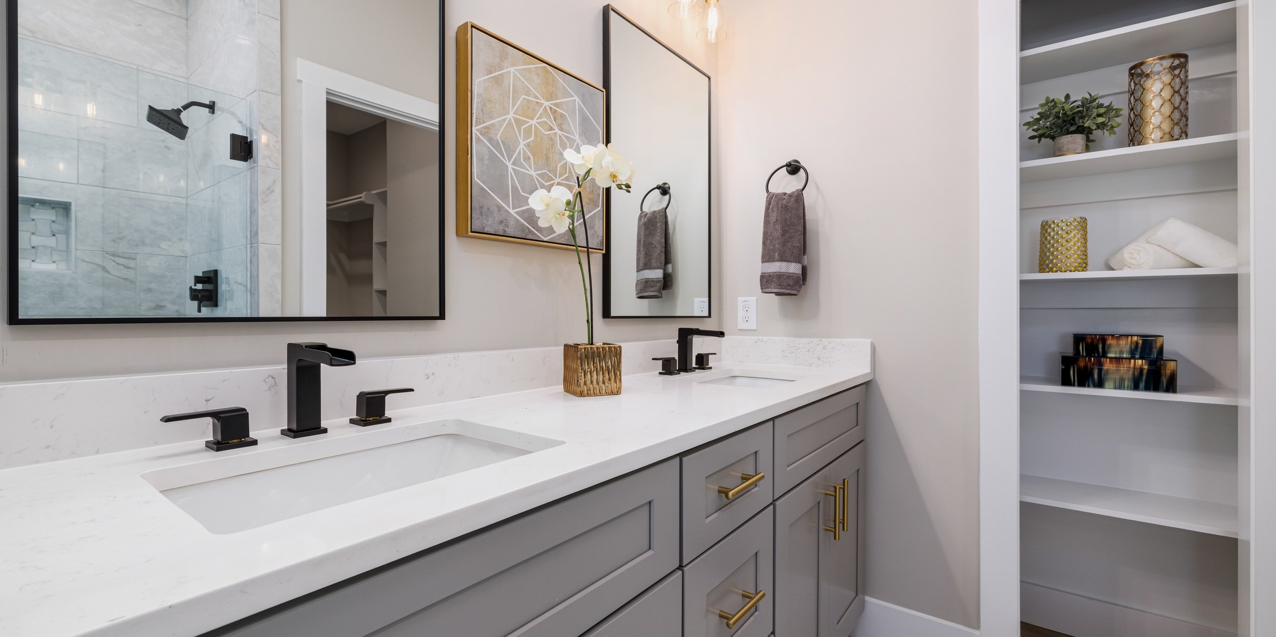 Modern bathroom vanity with gray cabinets, white stone countertop, two black faucets, large mirror, a flower vase, and artwork. Open shelves on the side with decorative items, and a glimpse of a shower with glass door in the reflection.