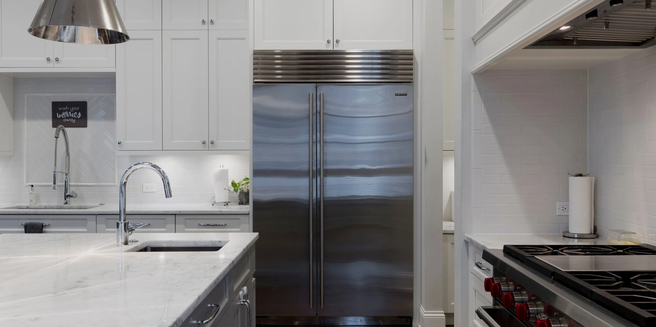Modern kitchen with white cabinets, stainless steel refrigerator, and a marble countertop island with dual sinks and chrome faucets. To the right is a stainless steel stove with red knobs and a white paper towel on a paper towel holder. There's a small black sign on the wall near the sink that reads "wash your worries away."