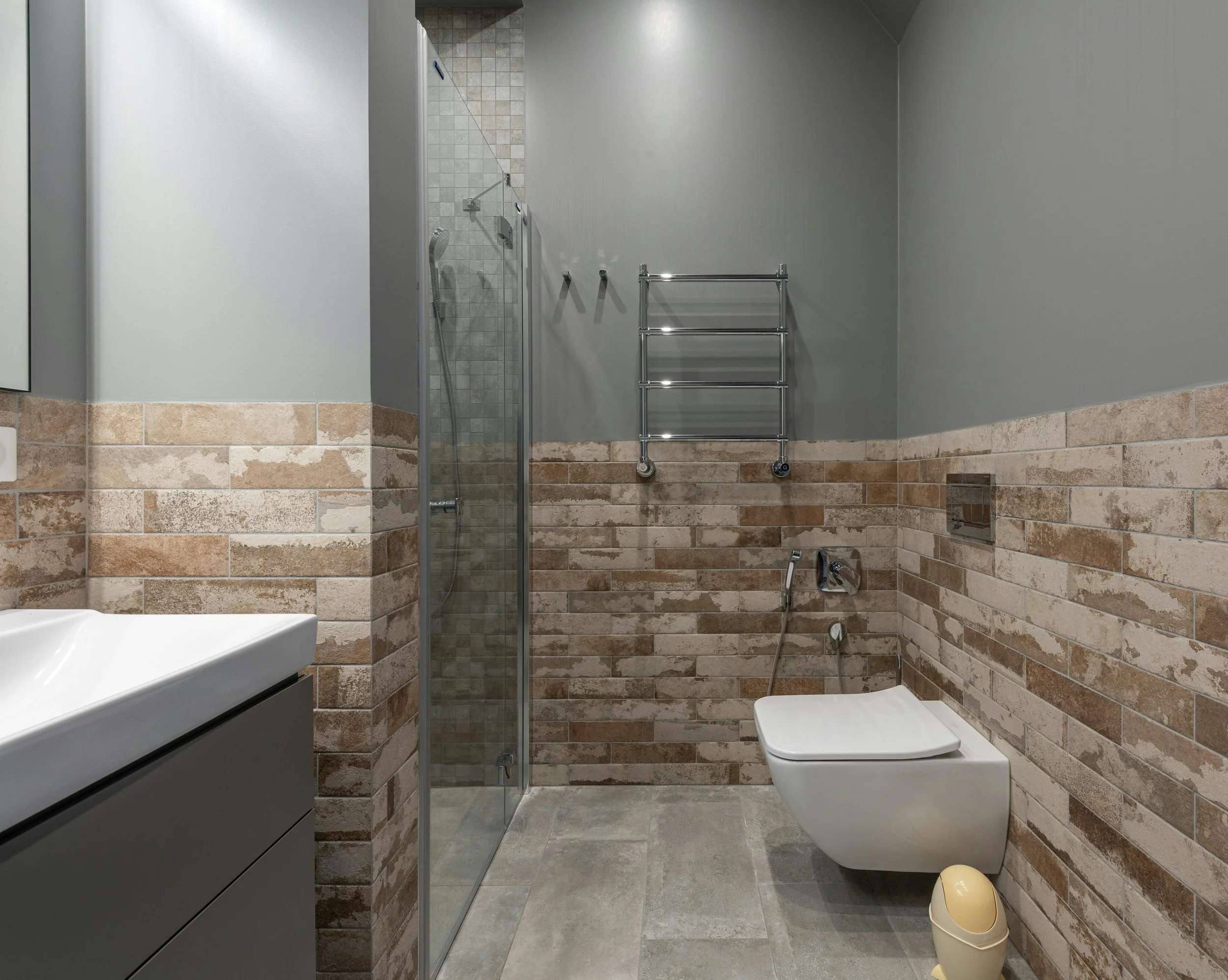 A modern bathroom with gray walls and brick-patterned tiles on the lower half. It features a glass-enclosed shower, a wall-mounted toilet, a gray vanity with a white sink, and a small yellow trash bin on the floor.