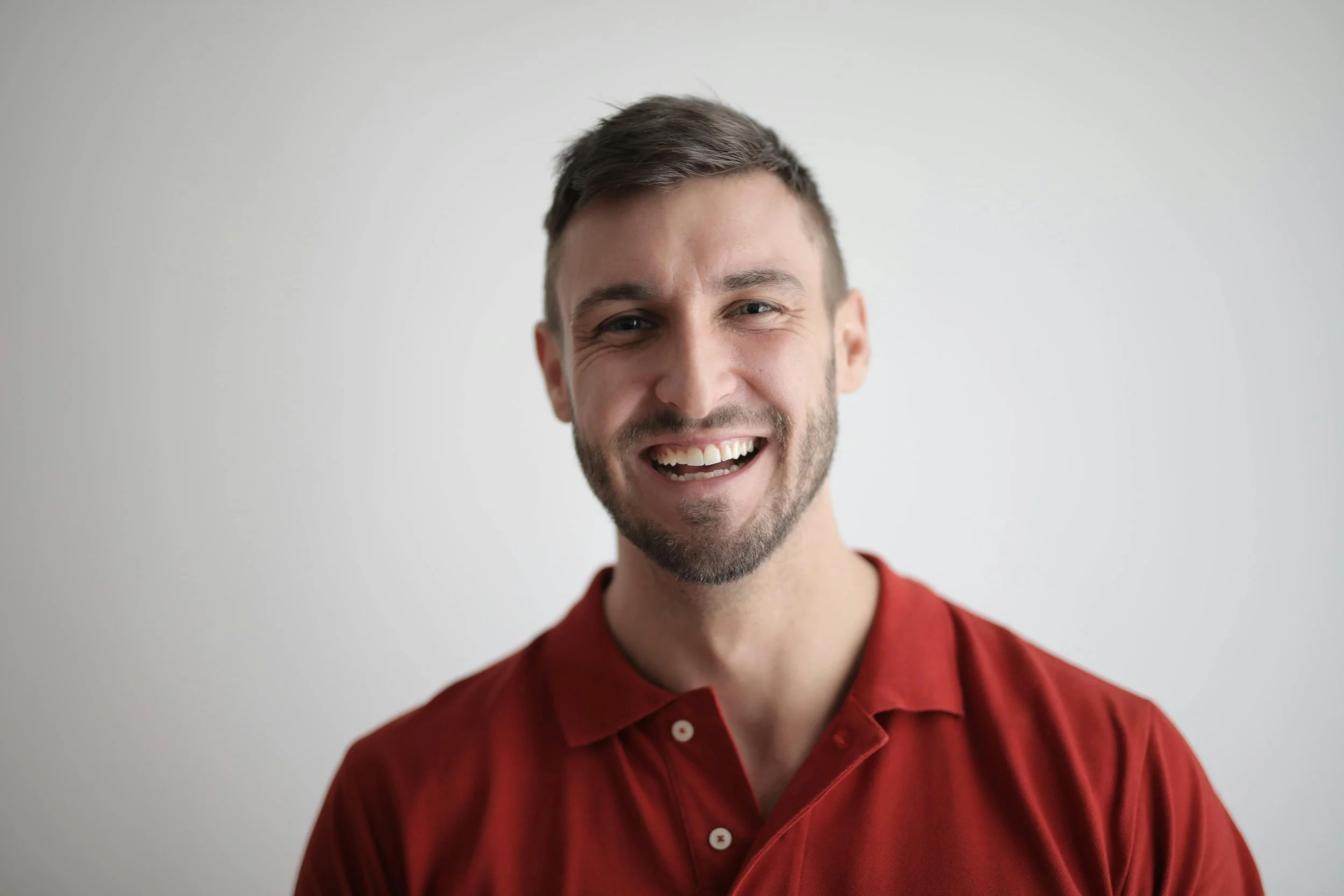 A smiling man with a short beard and brown hair wearing a red collared shirt against a plain light background.