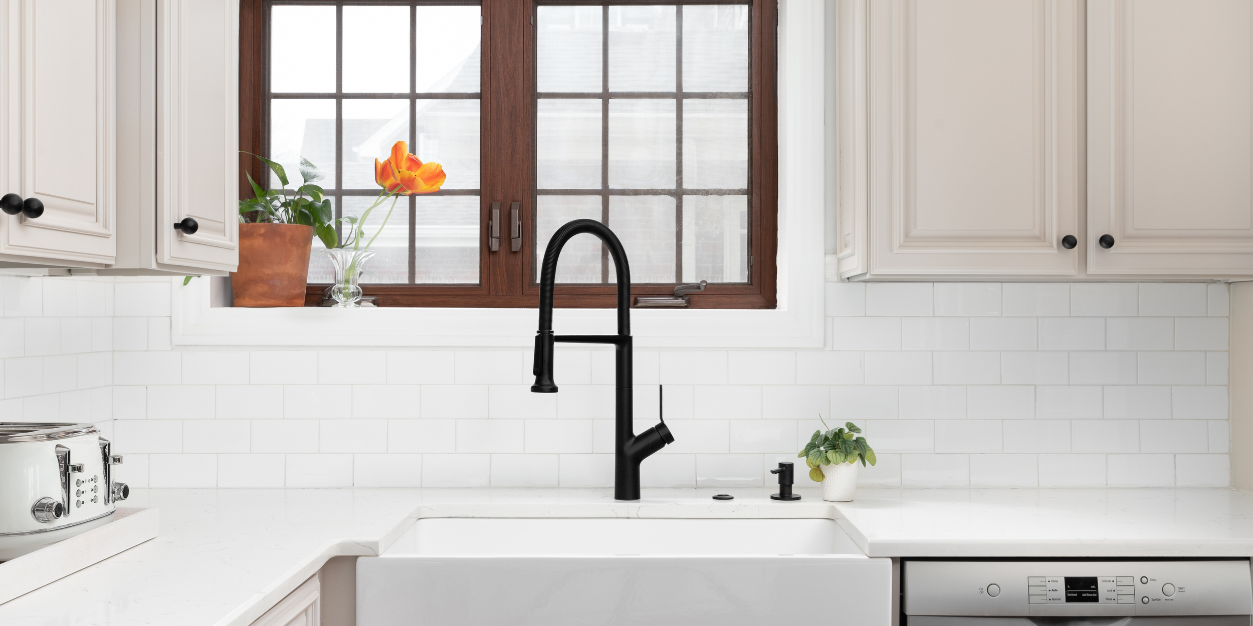 Kitchen sink with black faucet, window above, white cabinets, potted orange and green plant, and a small green plant in a white pot on the right.