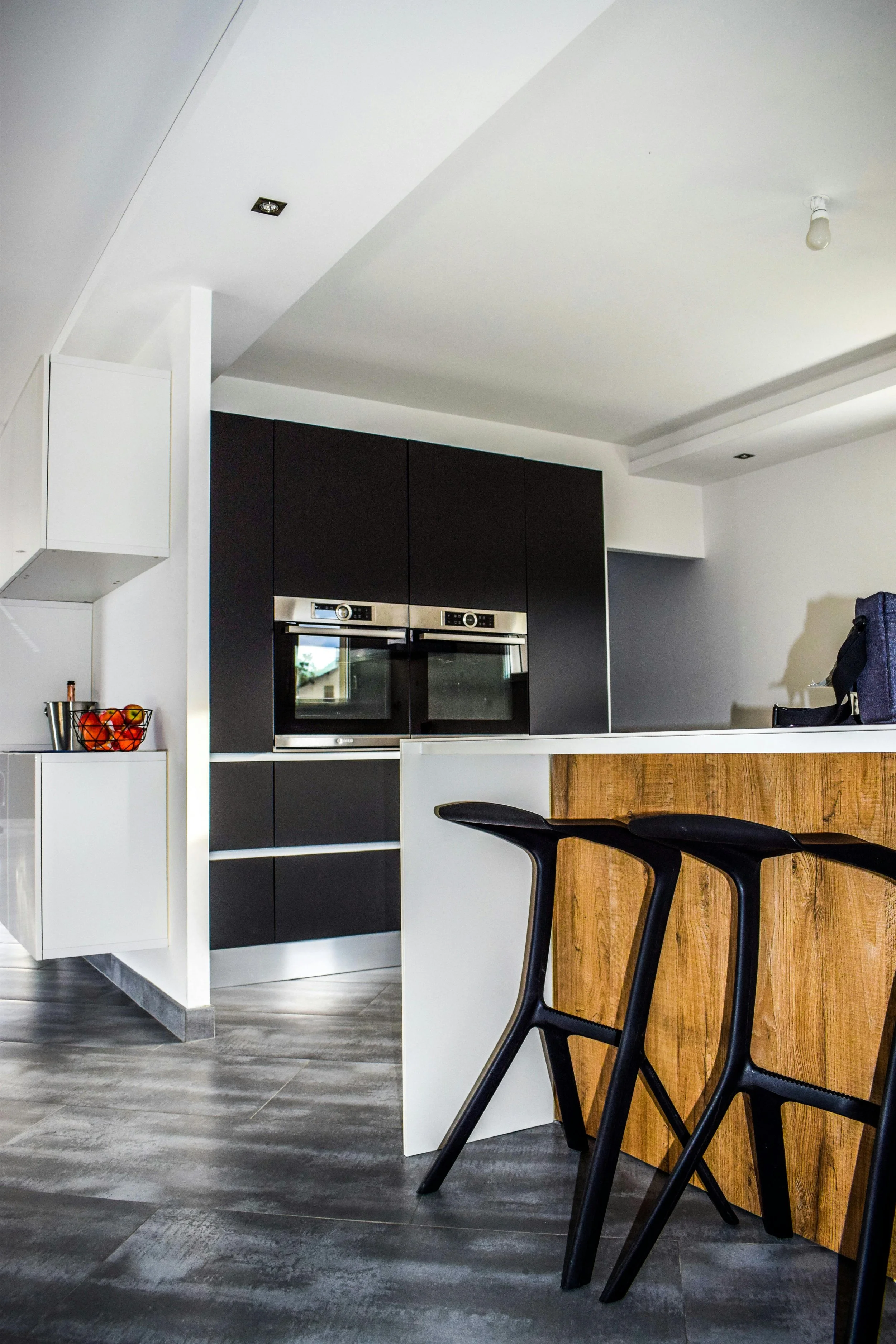 Modern kitchen with black cabinets, built-in ovens, a wooden breakfast bar, black chairs, and a gray tiled floor.
