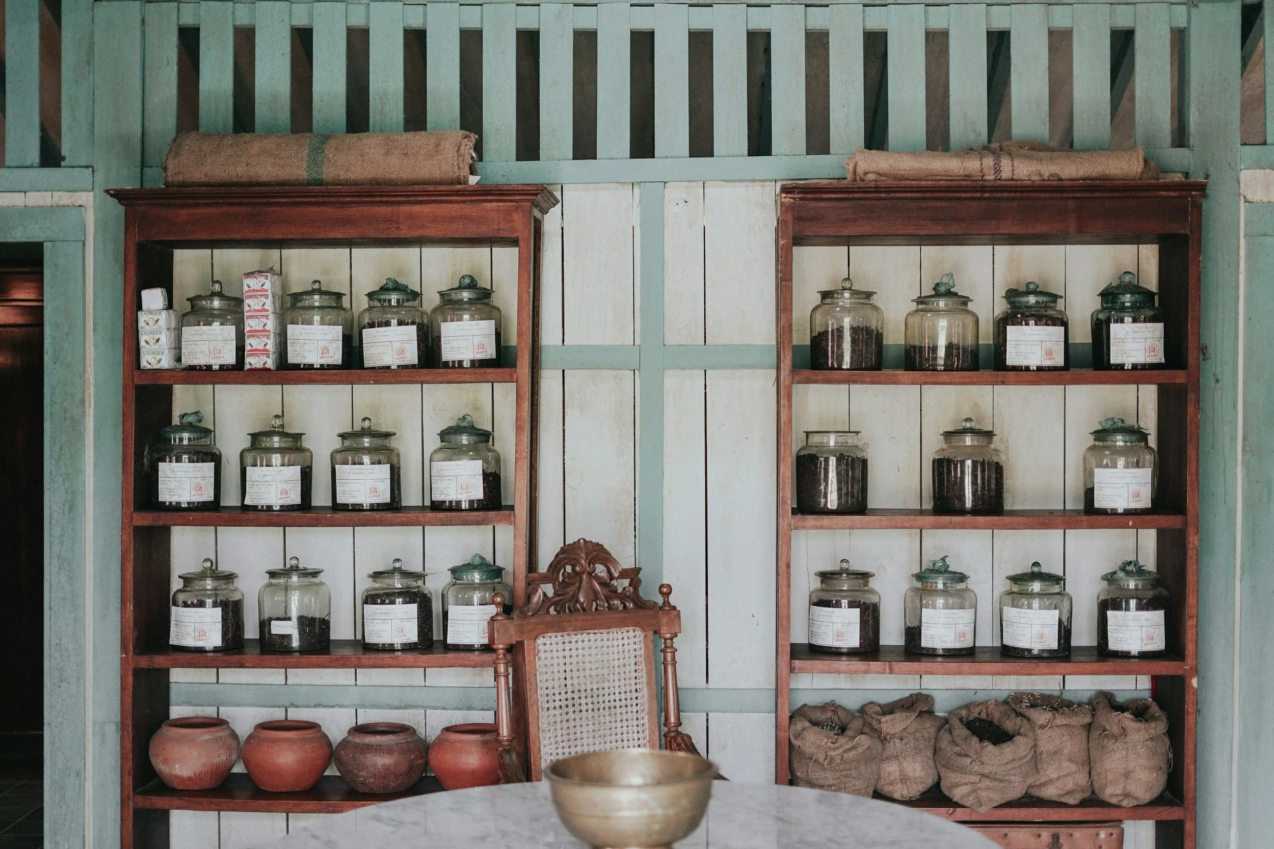 Two wooden shelves filled with glass jars containing herbs or spices, with name tags on each jar, placed against a wall with light teal and white vertical panels, with some burlap sacks on top of the shelves, a carved wooden chair with a woven cane backrest, and a marble-topped table in front.