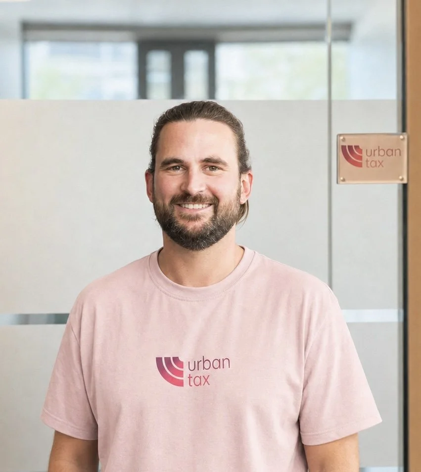 Ein lächelnder Mann in einem schwarzen Anzug und weißem T-Shirt vor einem hellen Hintergrund.