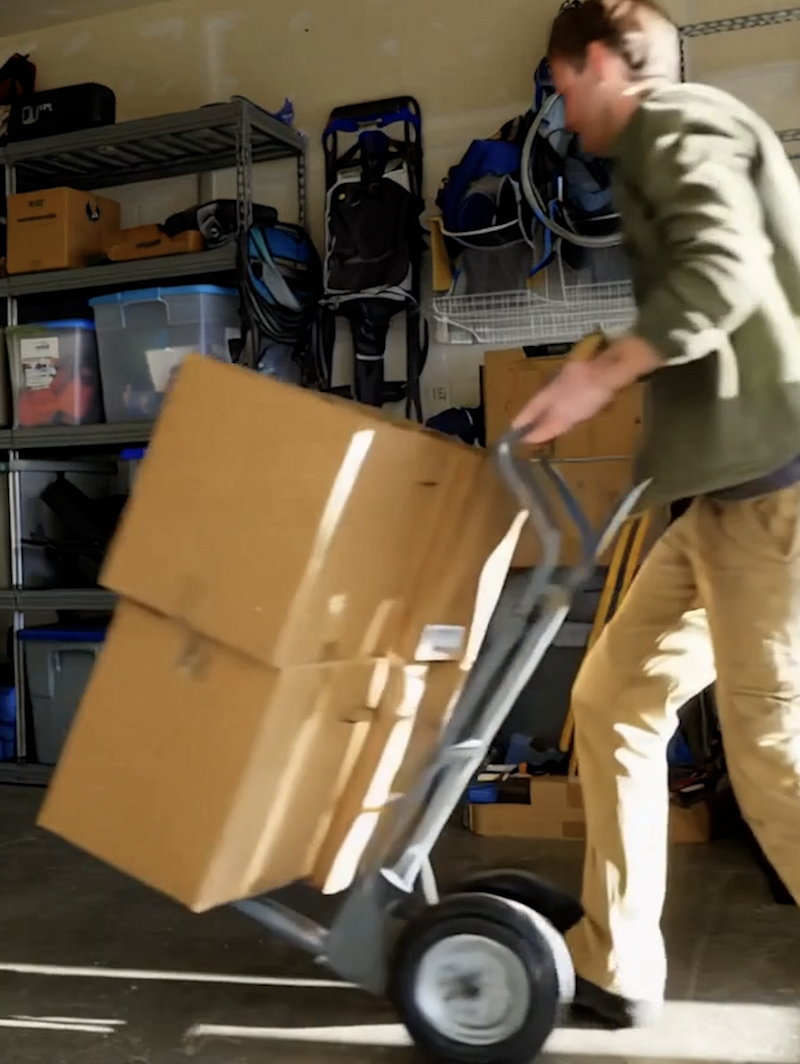 Person in a garage unloading or transporting boxes on a hand truck, with shelves and tools in the background.