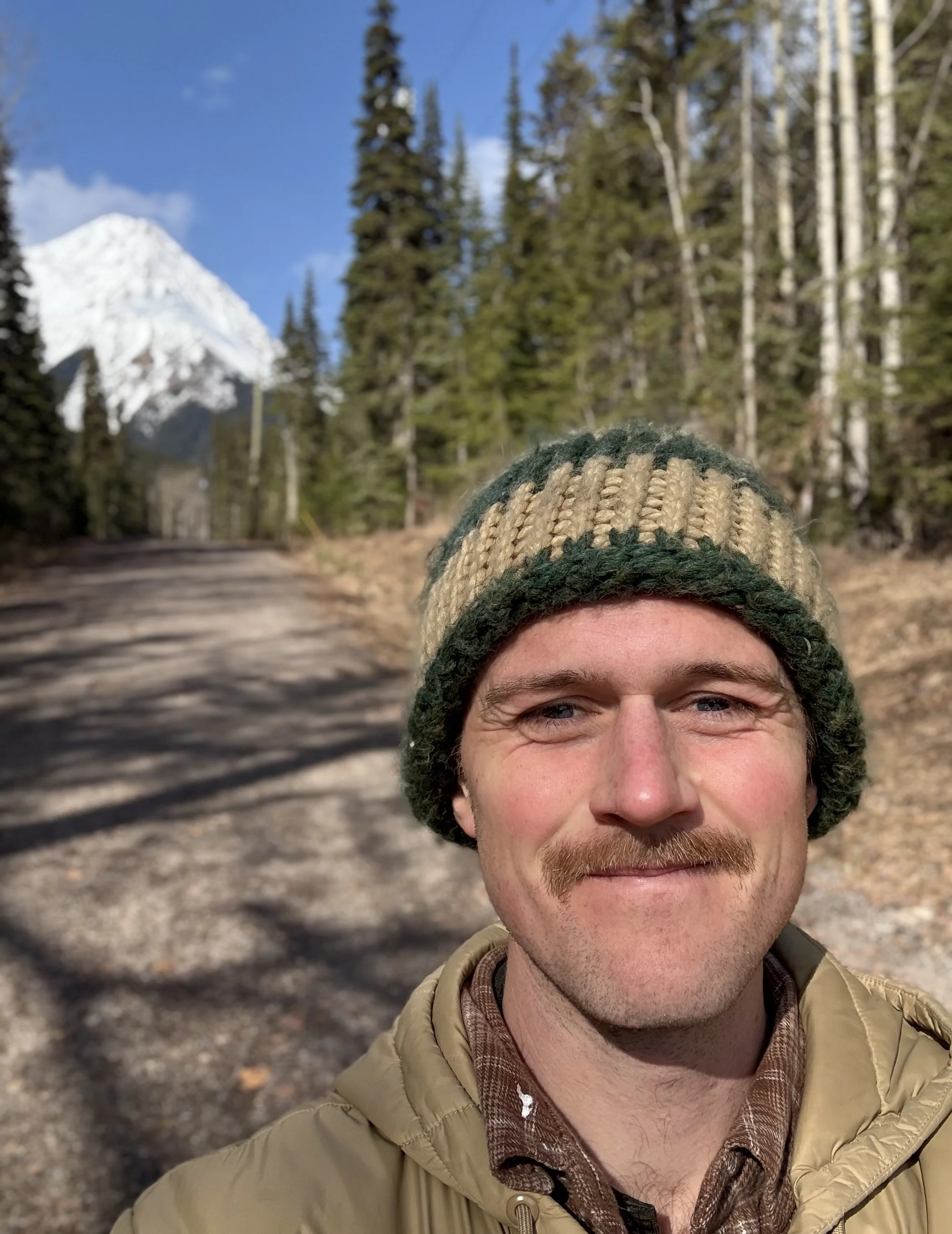 A man with a mustache and light facial hair wearing a green and tan knit beanie and a tan jacket, taking a selfie outdoors on a sunny day with a forest trail, tall trees, and a snow-capped mountain in the background.