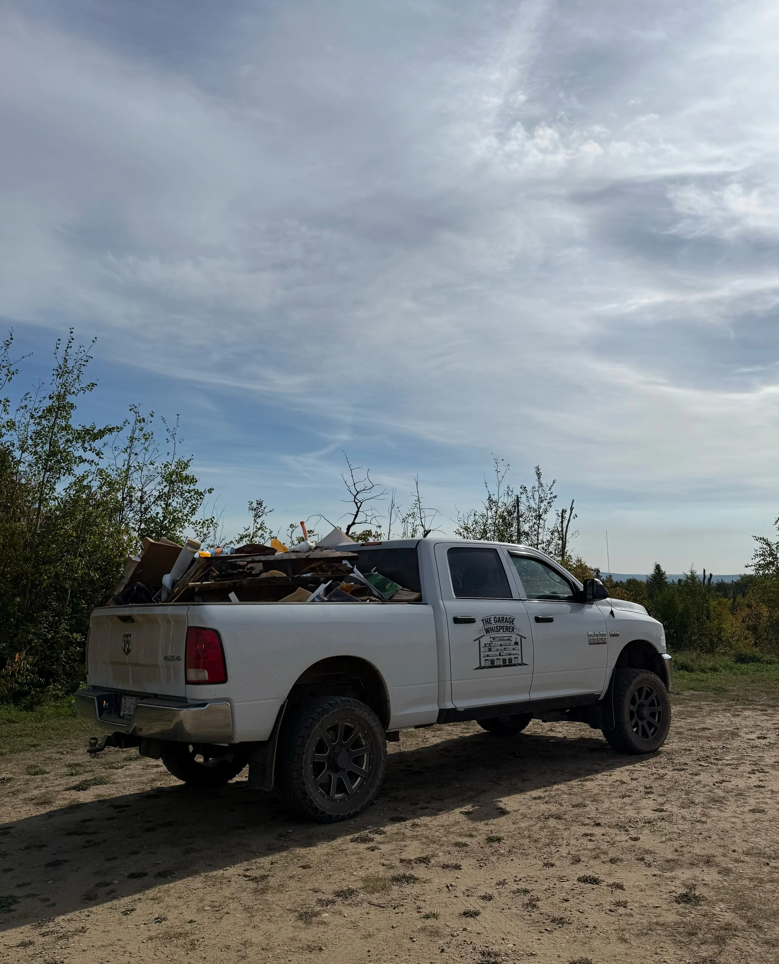 White pickup truck loaded with garbage and miscellaneous items parked outdoors on a dirt road under a cloudy sky.