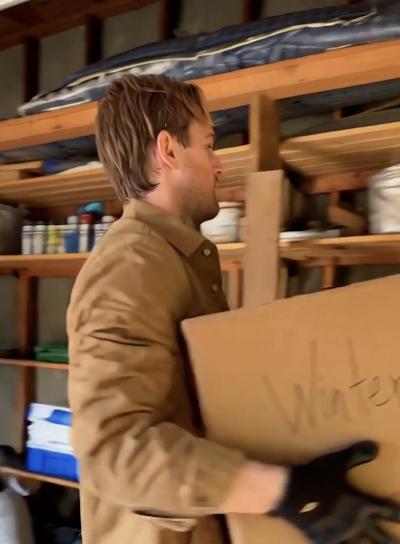 A man wearing a tan jacket and gloves is holding a cardboard box in a garage with wooden shelves. The shelves contain various cans, containers, and a blue cooler. A mattress is on a top bunk in the background.