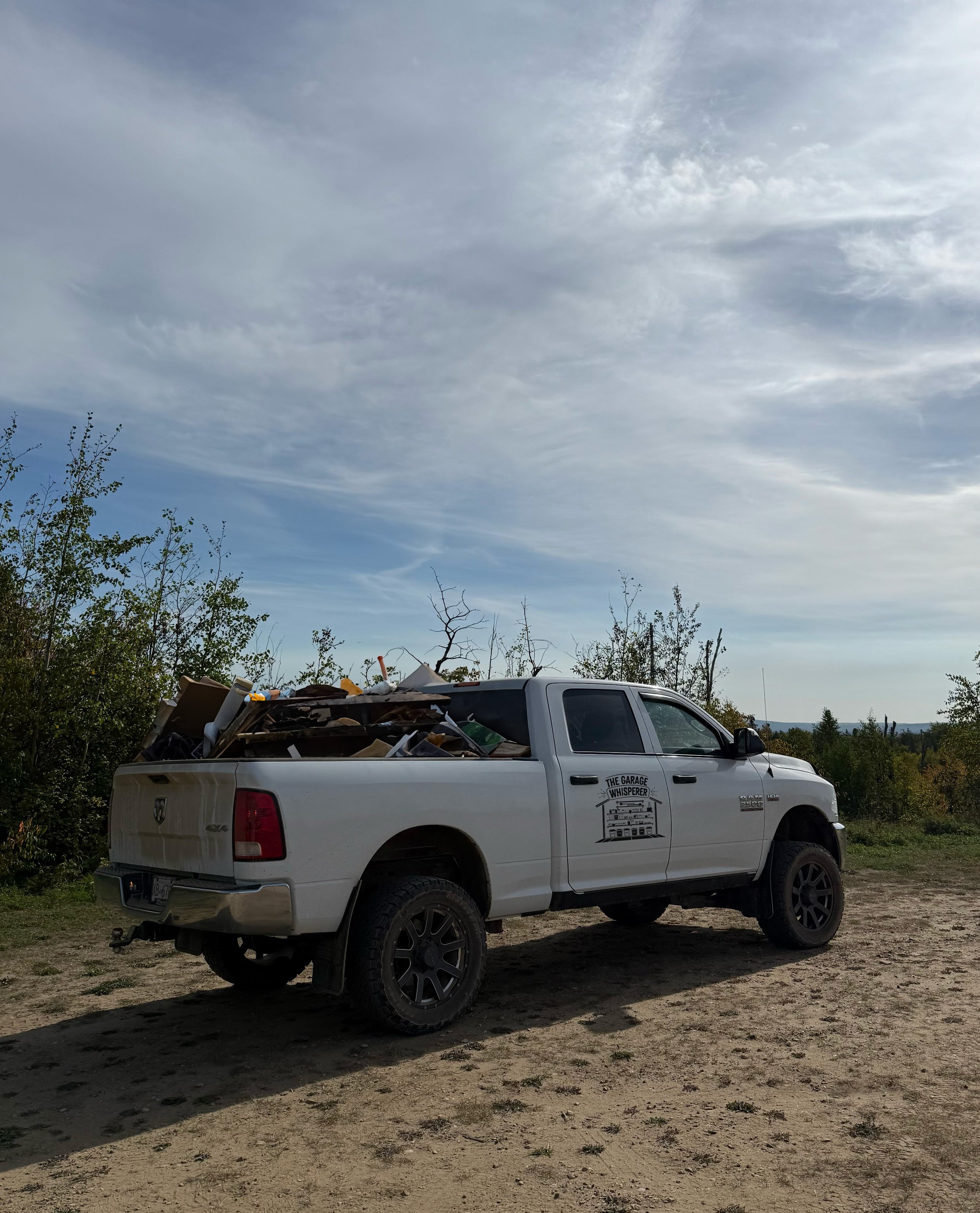 White pickup truck labeled 'The Garage Whisperer' parked outdoors on a dirt surface, loaded with garbage, with trees and a cloudy sky in the background.