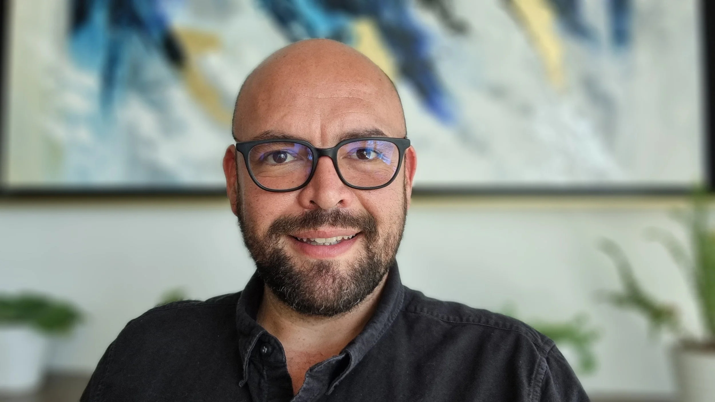 Hombre calvo con barba, gafas y camisa negra sonriendo en oficina con arte abstracto y plantas de fondo.