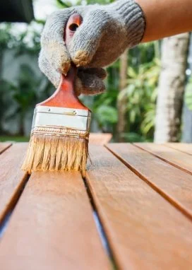 Hand wearing a glove using a brush to stain a wooden surface outdoors.