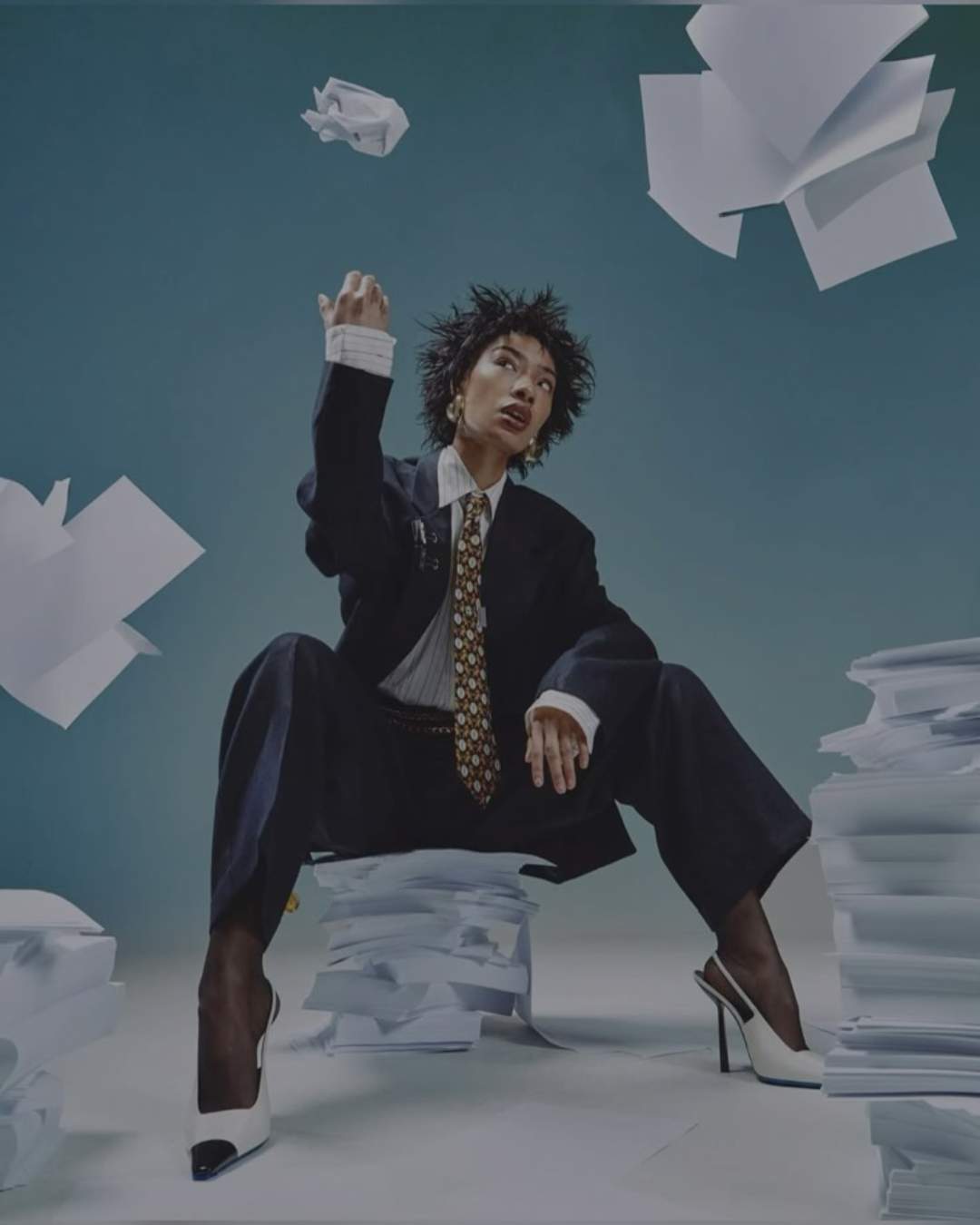 A woman dressed in a black suit, sitting among stacks of papers and surrounded by flying sheets, wearing high heels and a patterned tie, with a confident expression. She uses WeShortlist's AI candidate screening software