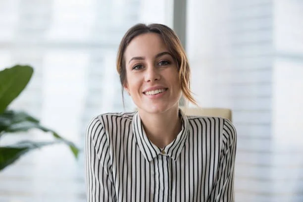 Smiling woman in a striped shirt sitting in an office with a large window and a potted plant in the background.