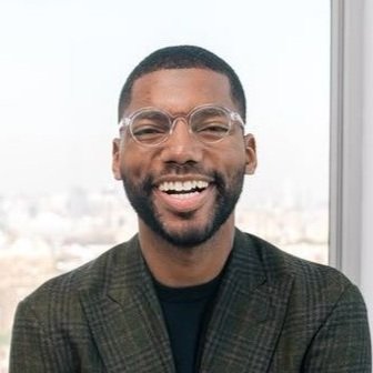 Smiling man with glasses and a beard wearing a green blazer in front of a window