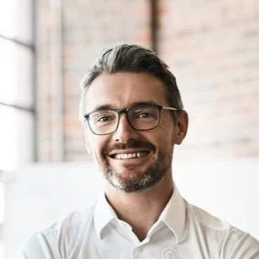 Portrait of a smiling man with glasses, dark hair, and a beard, wearing a white shirt, standing indoors near a brick wall window.