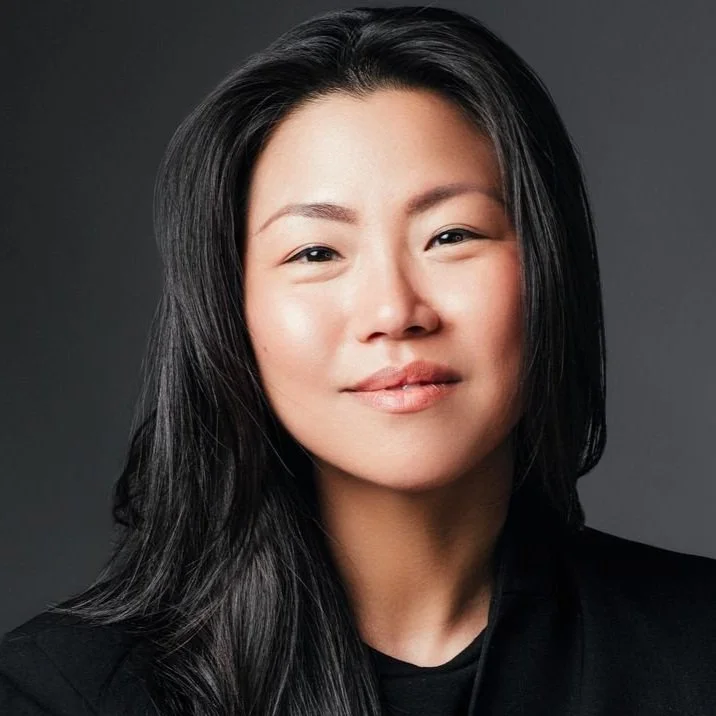 Close-up portrait of an Asian woman with long black hair, subtle smile, wearing a black top, against a dark gray background.