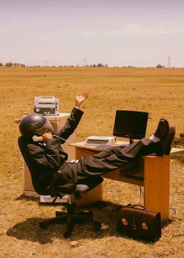 A man in a business suit, wearing a motorcycle helmet, is sitting at an office desk in a barren field. He is talking on a landline phone, with one arm raised, and resting his feet on the desk. The desk holds a computer monitor, keyboard, and a typewr