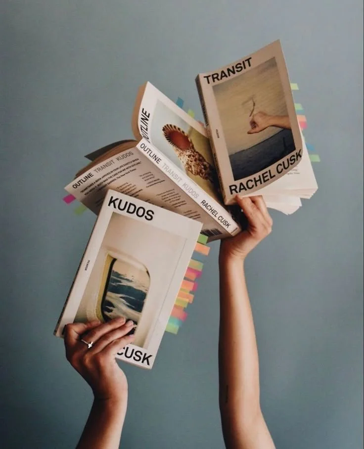 A person holding three books titled "Kudos," "Transit," and "Outline Transit Kudus" by Rachel Cusk. The books have colorful sticky tabs along their edges, and the background is a plain blue wall.