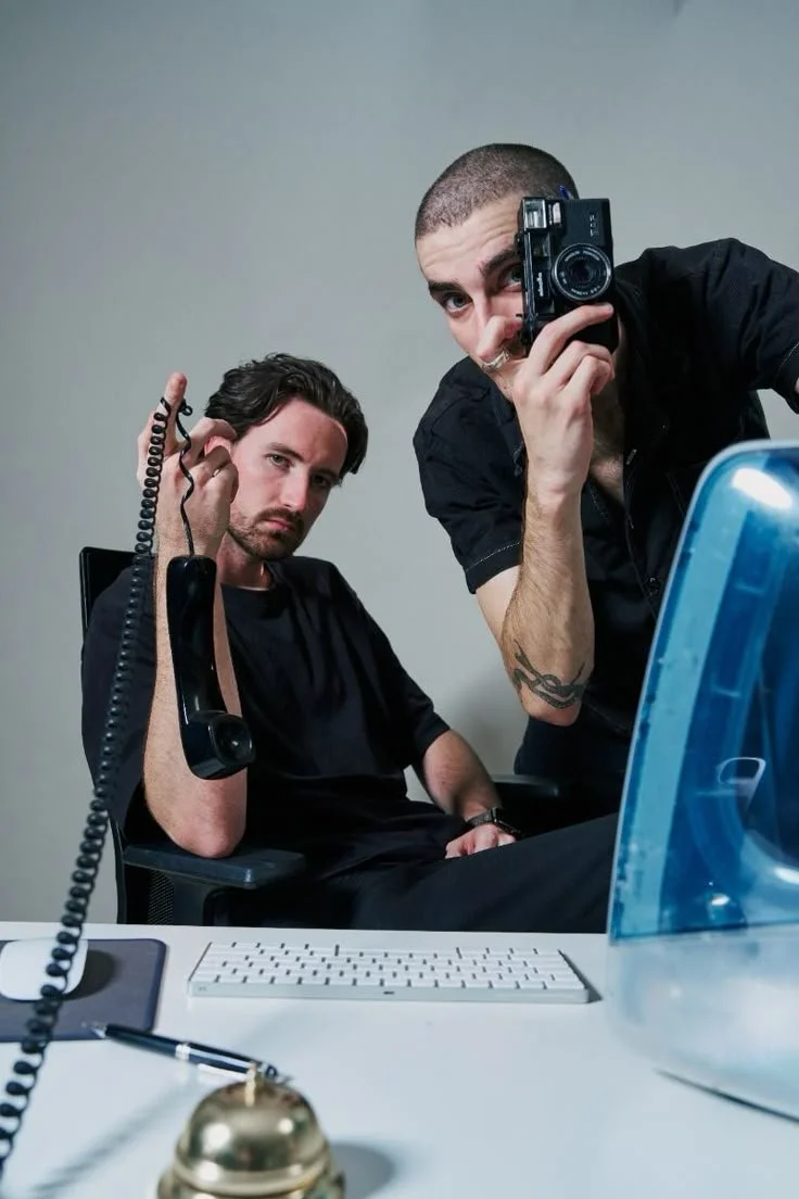 Two young men in black shirts taking a selfie in an office setting, one holding a camera and the other holding a telephone, with office supplies on the desk.
