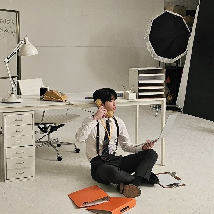 A young man in office attire sitting on the floor in a photography studio, holding a vintage rotary phone to his ear with one hand and a laptop in the other. The studio background includes a white desk, a white office chair, a large studio umbrella, 