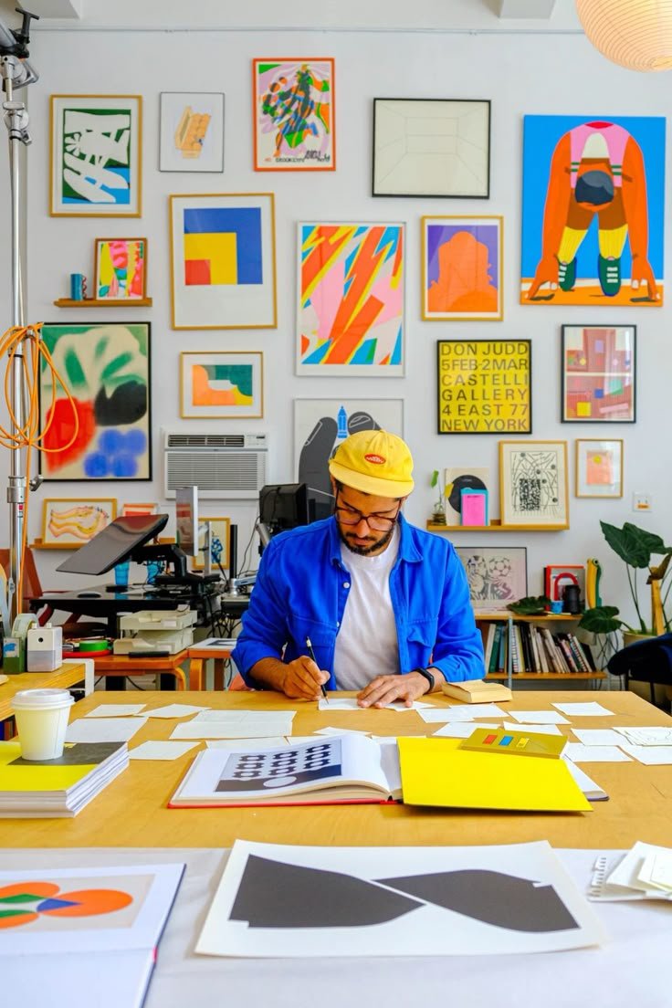 A man with glasses and a beard wearing a yellow cap and a blue jacket sitting at a cluttered desk with papers around him, in an art studio with colorful abstract artwork on the walls.