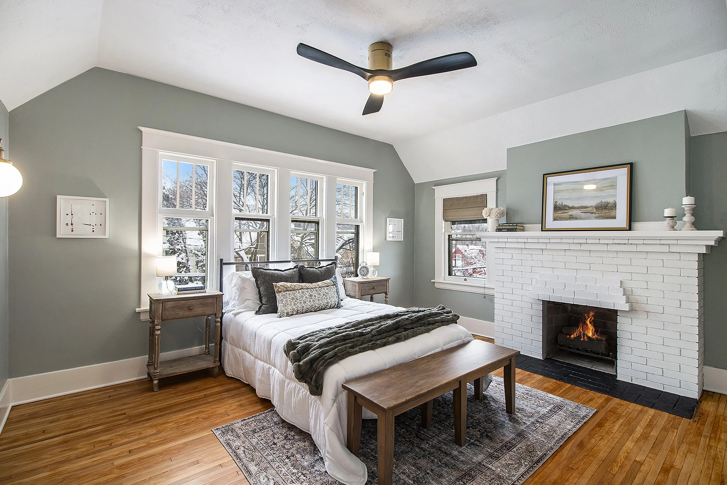 A cozy bedroom with light green walls, a white brick fireplace with a fire, a bed with white linens and decorative pillows, wooden nightstands, a ceiling fan, and multiple windows showing a snowy outdoor scene.