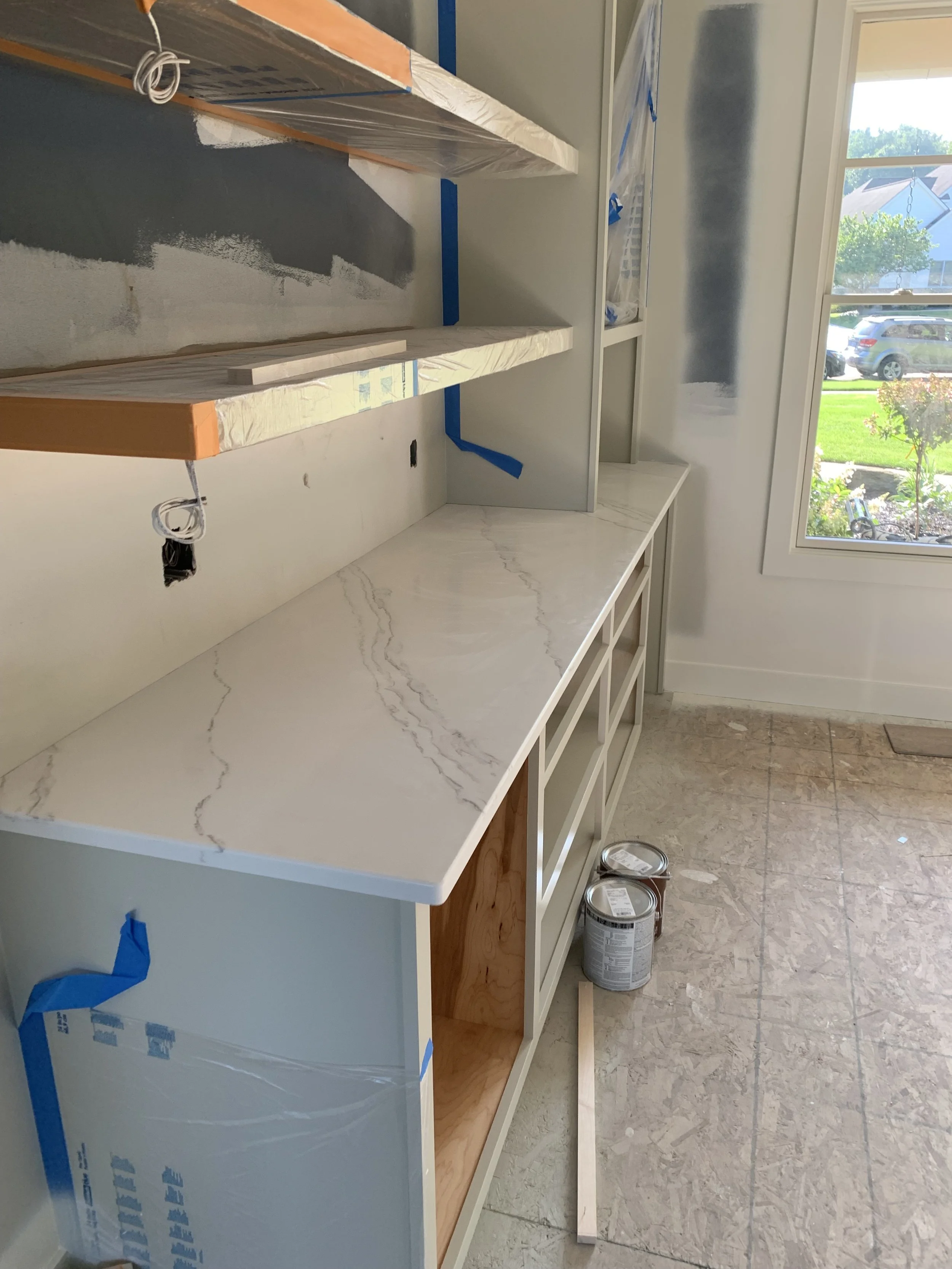 A kitchen or workspace under construction with a white marble countertop, open white cabinets, and wall shelves covered with plastic and painter's tape, near a window showing a sunny outdoor scene.