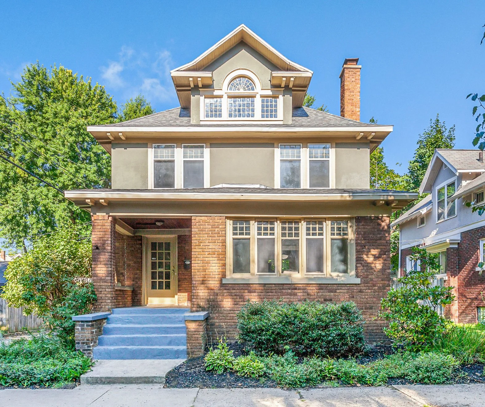 A three-story house with brick and beige siding, a covered front porch, large front windows, and landscaping with bushes and plants under a blue sky.