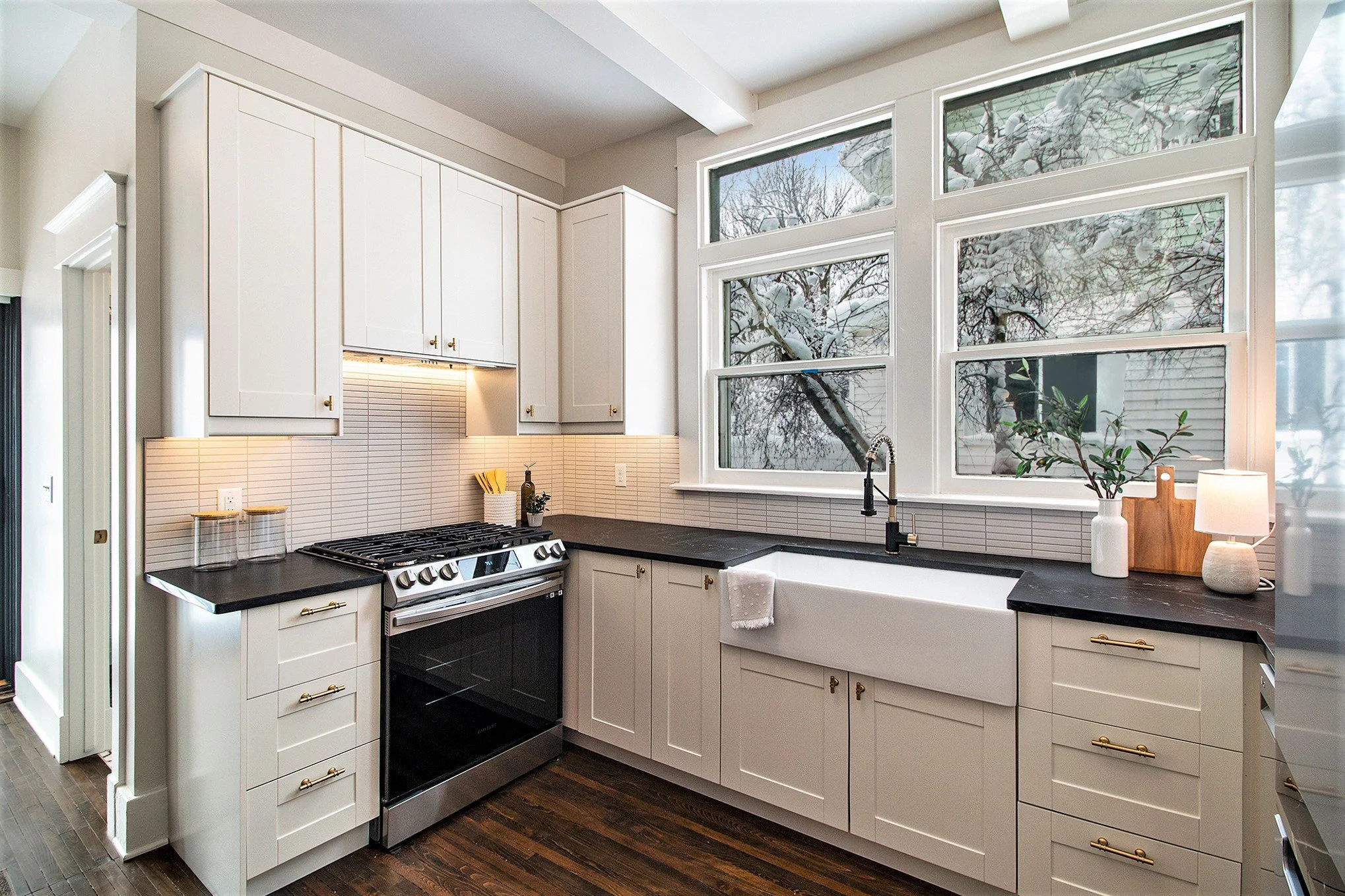 Modern kitchen with white cabinets, black countertops, and a farmhouse sink under large windows showing a snowy outdoor scene.