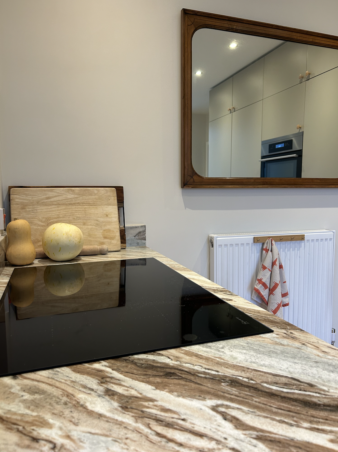 Kitchen countertop with a black electric cooktop, a wooden cutting board, and decorative gourds. There is a large mirror on the wall reflecting kitchen cabinets and an oven, and a towel hanging on a radiator.